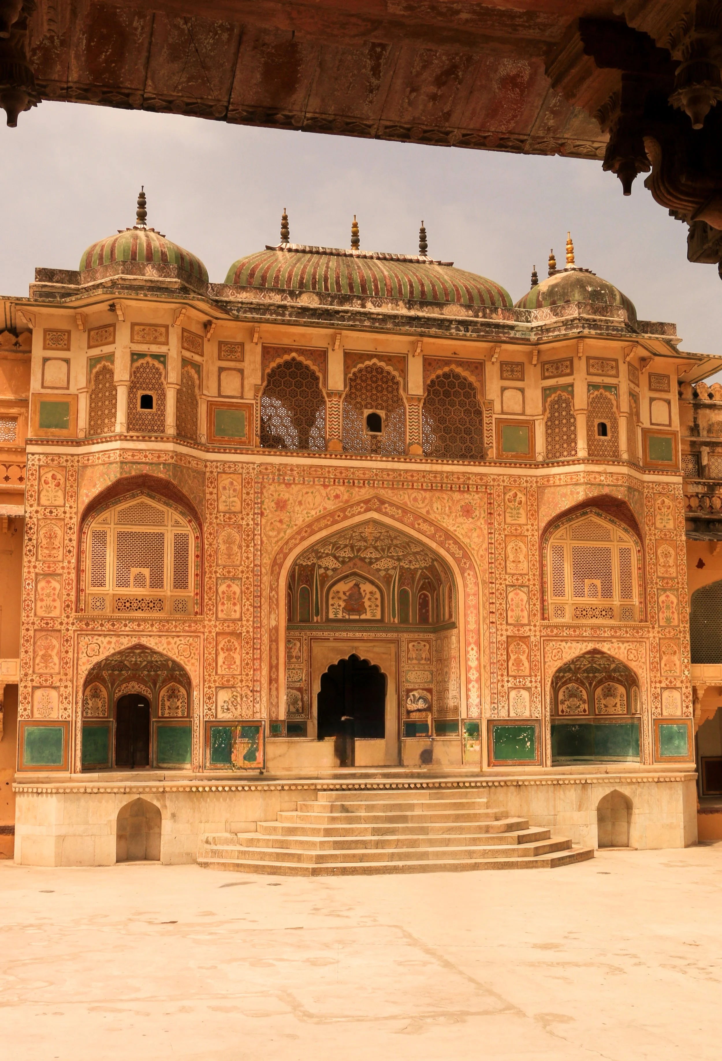 A historic Mughal palace featuring intricate tile work, arched windows, and ornate domes, with stairs leading up to the entrance, set against a cloudy sky.