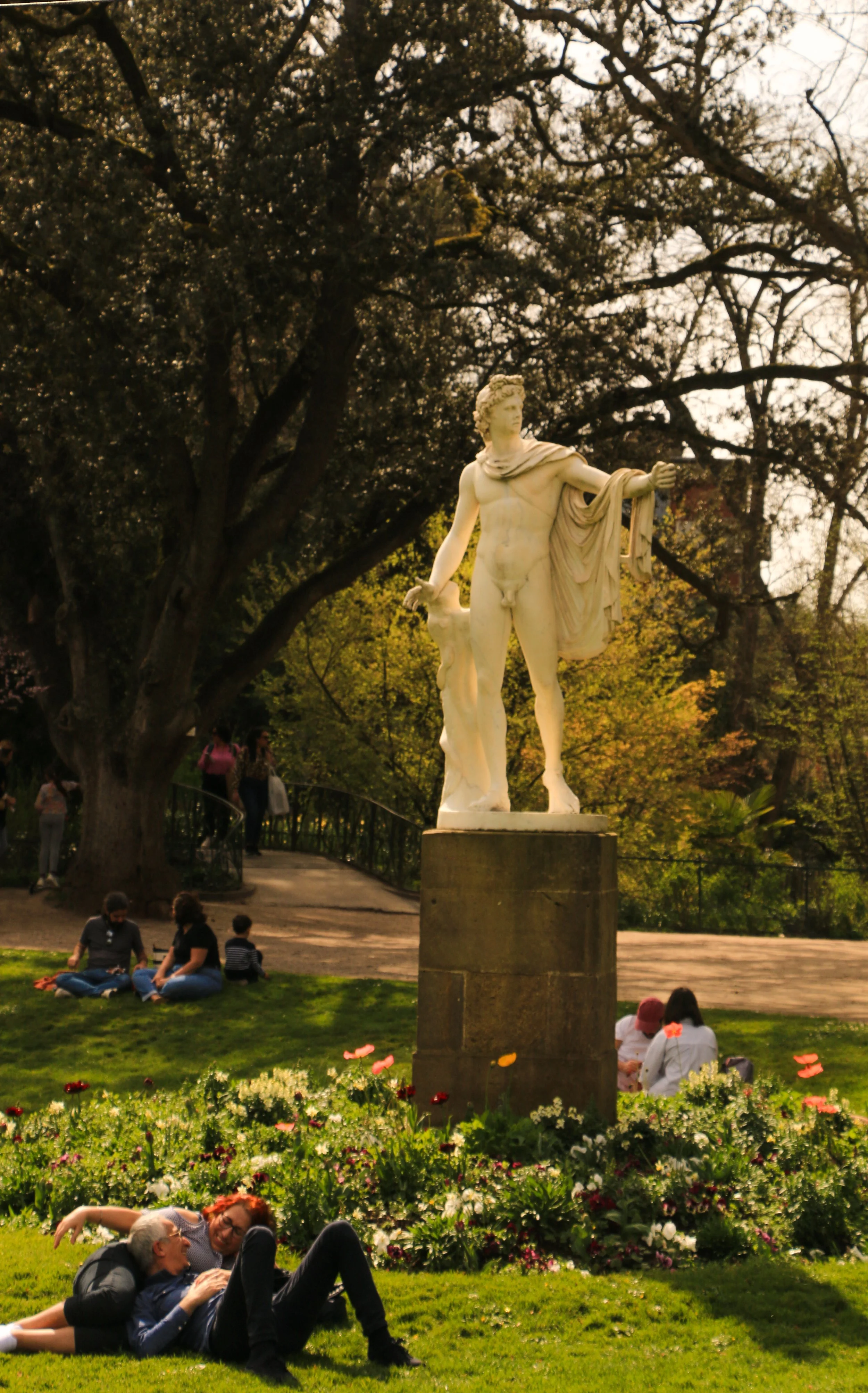 White marble statue of a male figure with a draped garment, standing outdoors in a park surrounded by trees, flowers, and people sitting and relaxing.