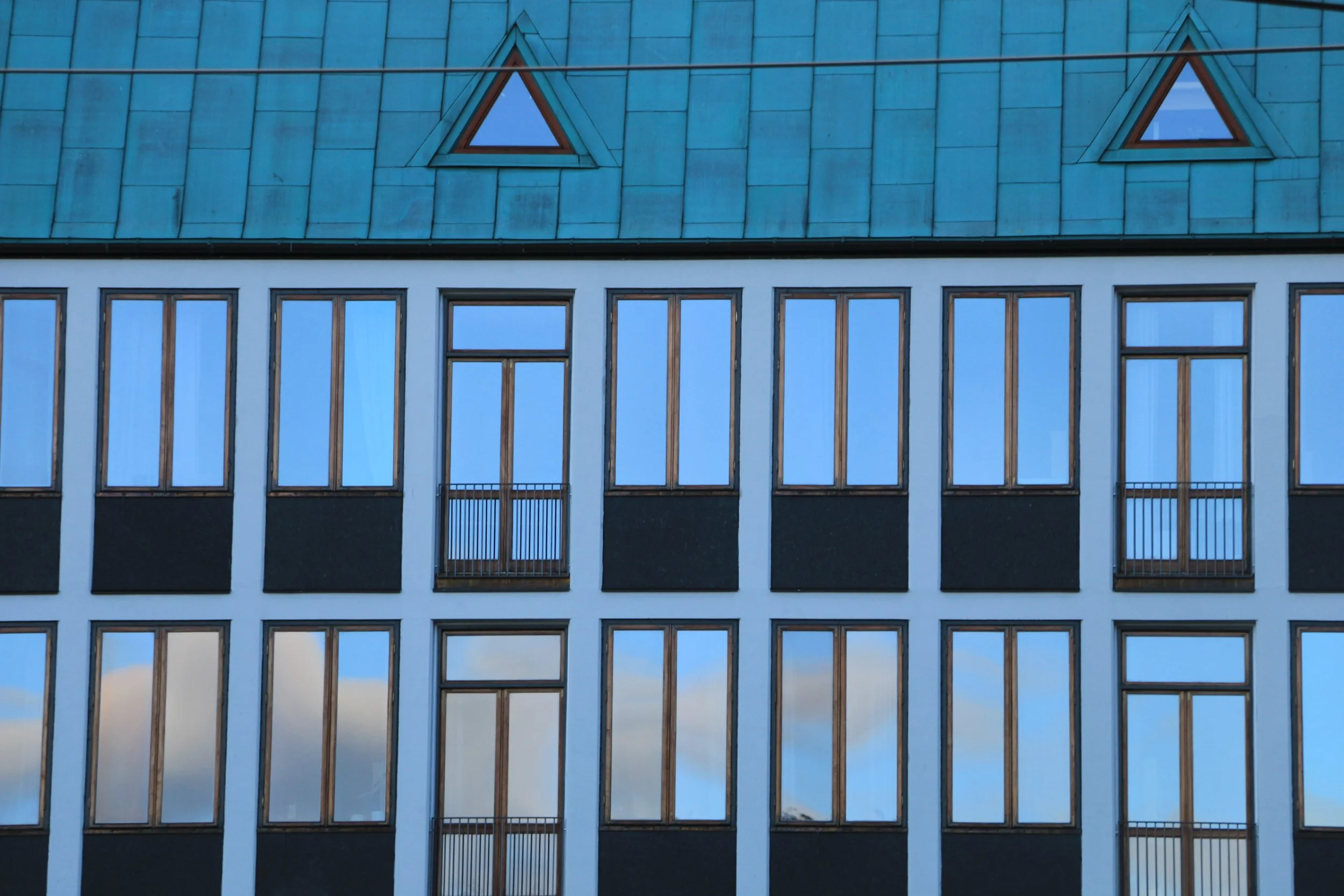 Modern multi-story building with large rectangular windows, some with small balconies, and a blue-tiled roof with triangular windows.