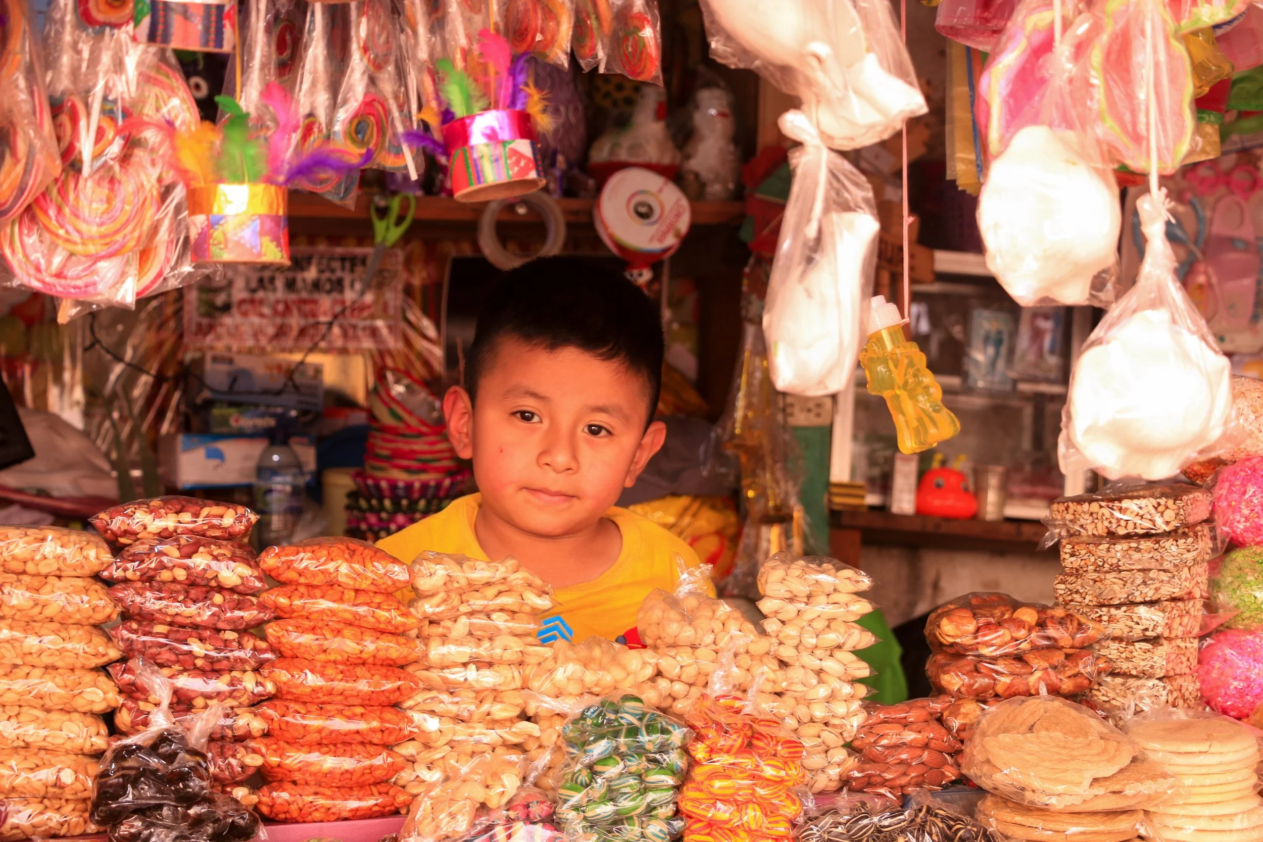 A young boy in a yellow shirt standing behind a stall filled with assorted packaged snacks, in a market with various colorful items hanging and displayed around him.