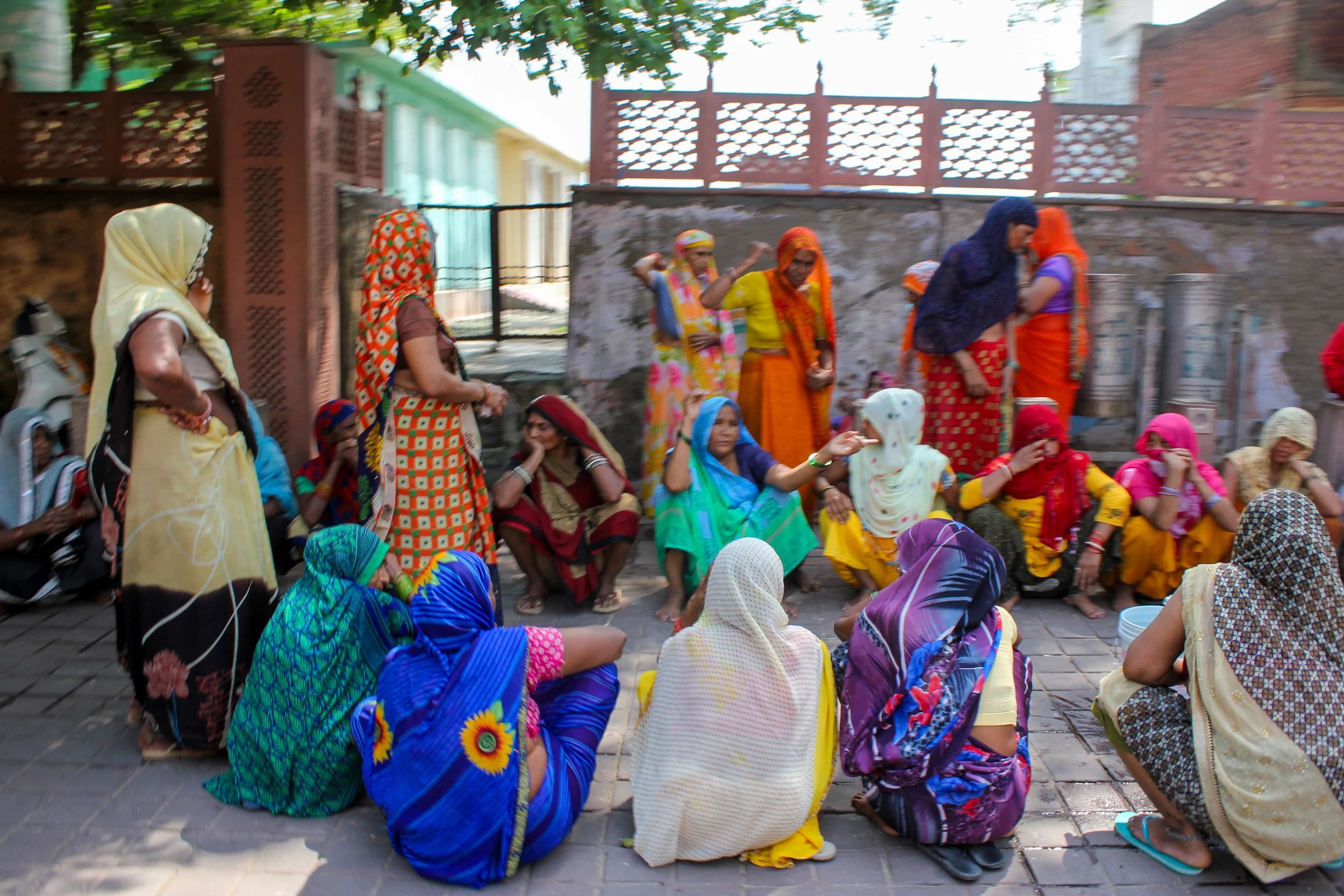 Group of women seated and standing outdoors, dressed in colorful traditional Indian attire, engaging in a conversation or meeting.