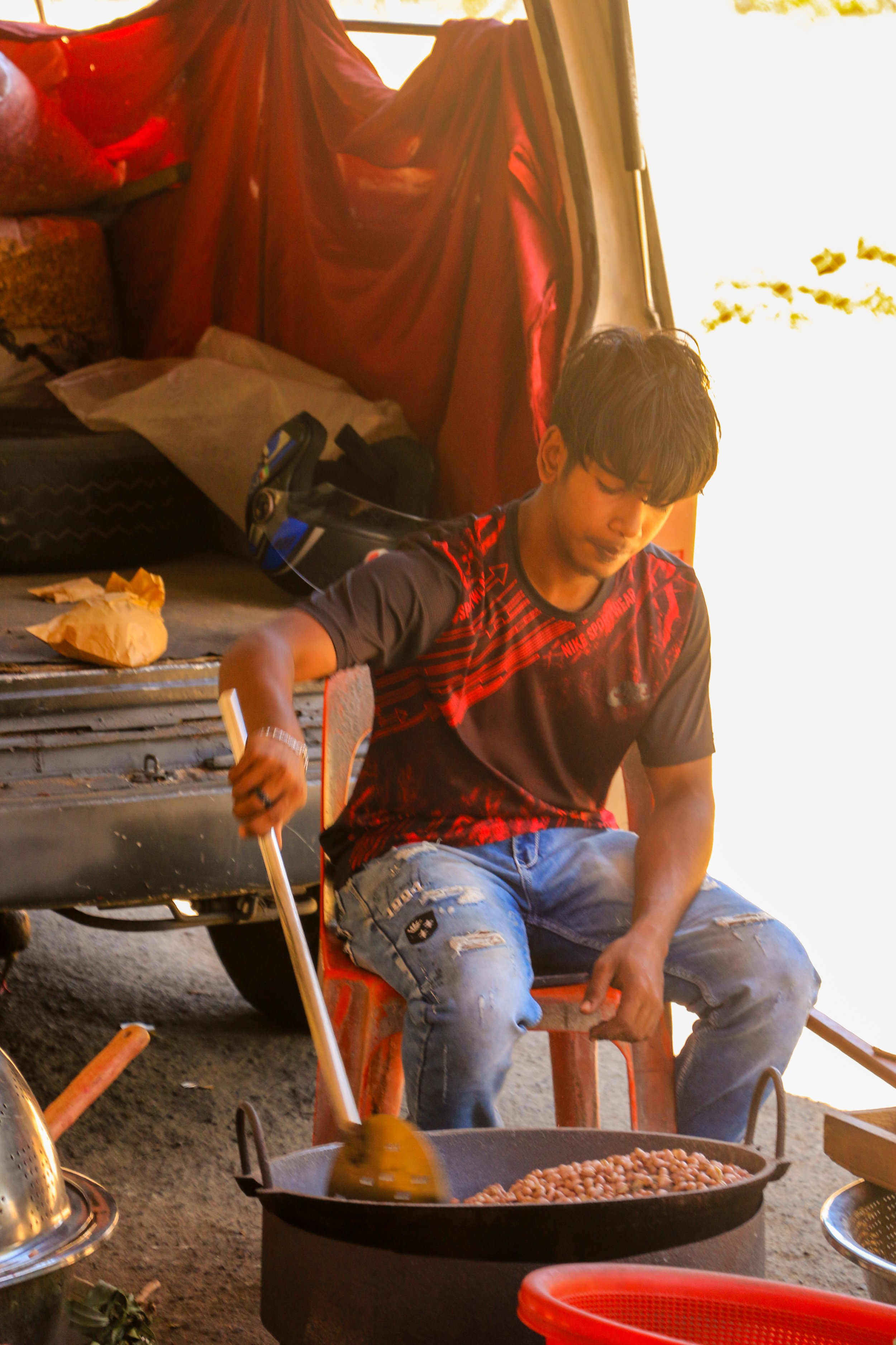 Young man cooking beans in a black pan over an open fire outdoors during daytime, with red draped fabric and various objects in the background.