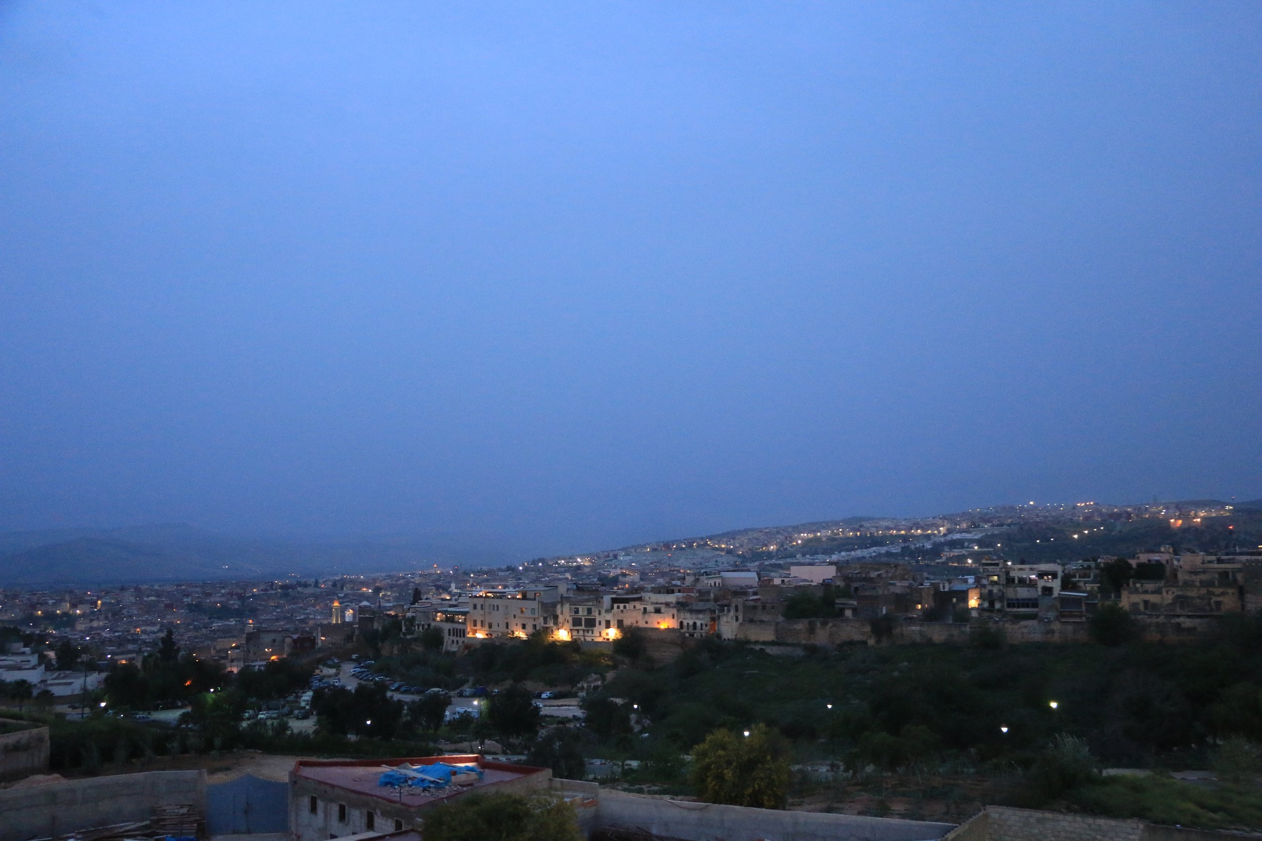 Cityscape at dusk with buildings illuminated, hills in the background, and a blue sky.