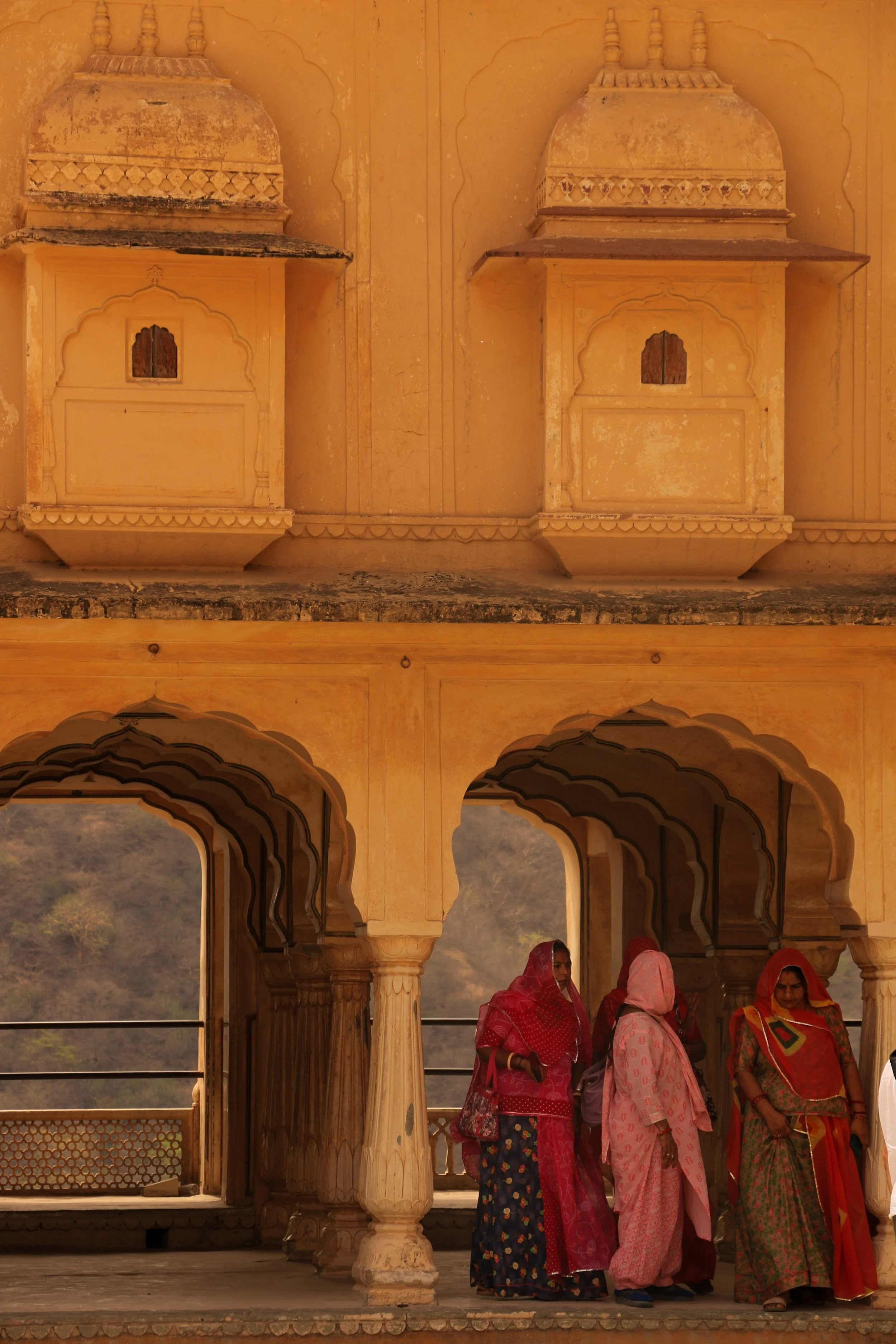 Women dressed in colorful traditional Indian attire walking inside a historic palace with ornate arches and balconies.