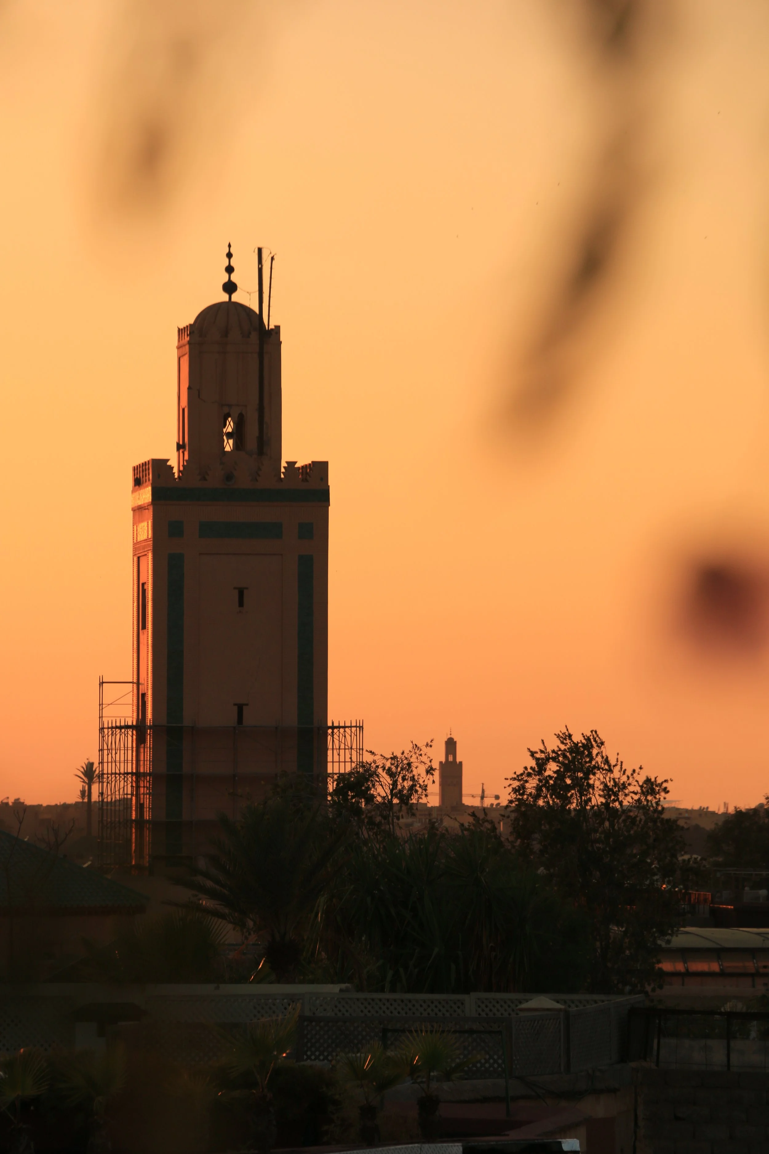 A silhouette of a tall, historic tower against a warm, orange sunset sky, with trees and buildings in the foreground.