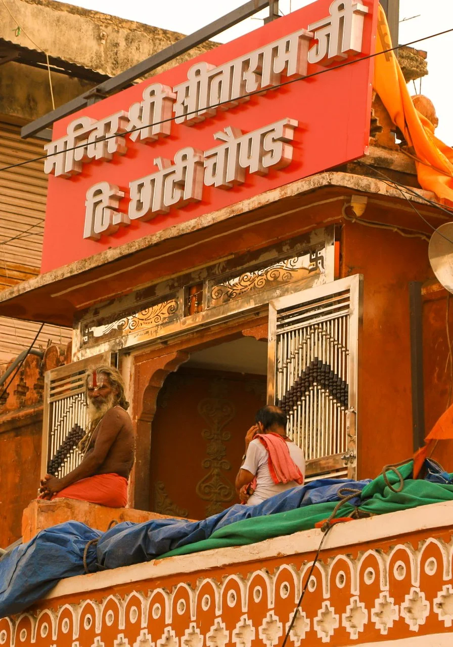 A building with a sign in Hindi and a man sitting on the balcony, with another person talking on a phone nearby.