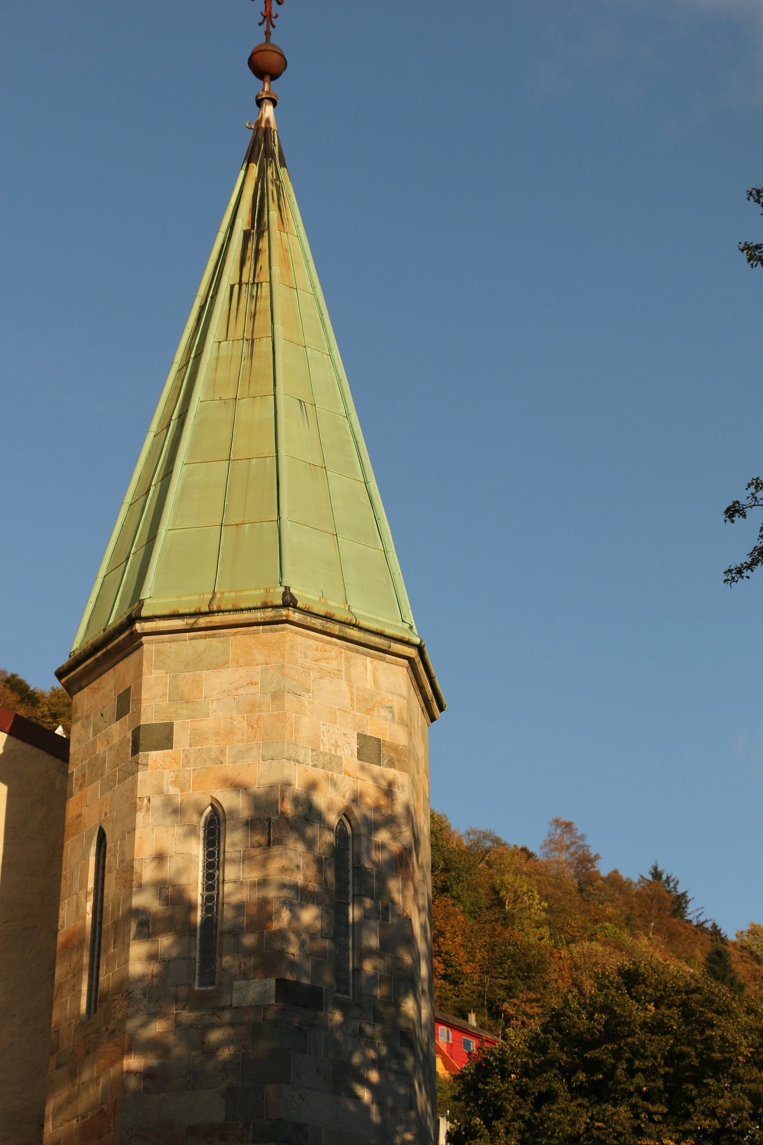 A church steeple with a green copper roof and stone base against a clear blue sky. Shadows from nearby trees are cast on the stone tower.