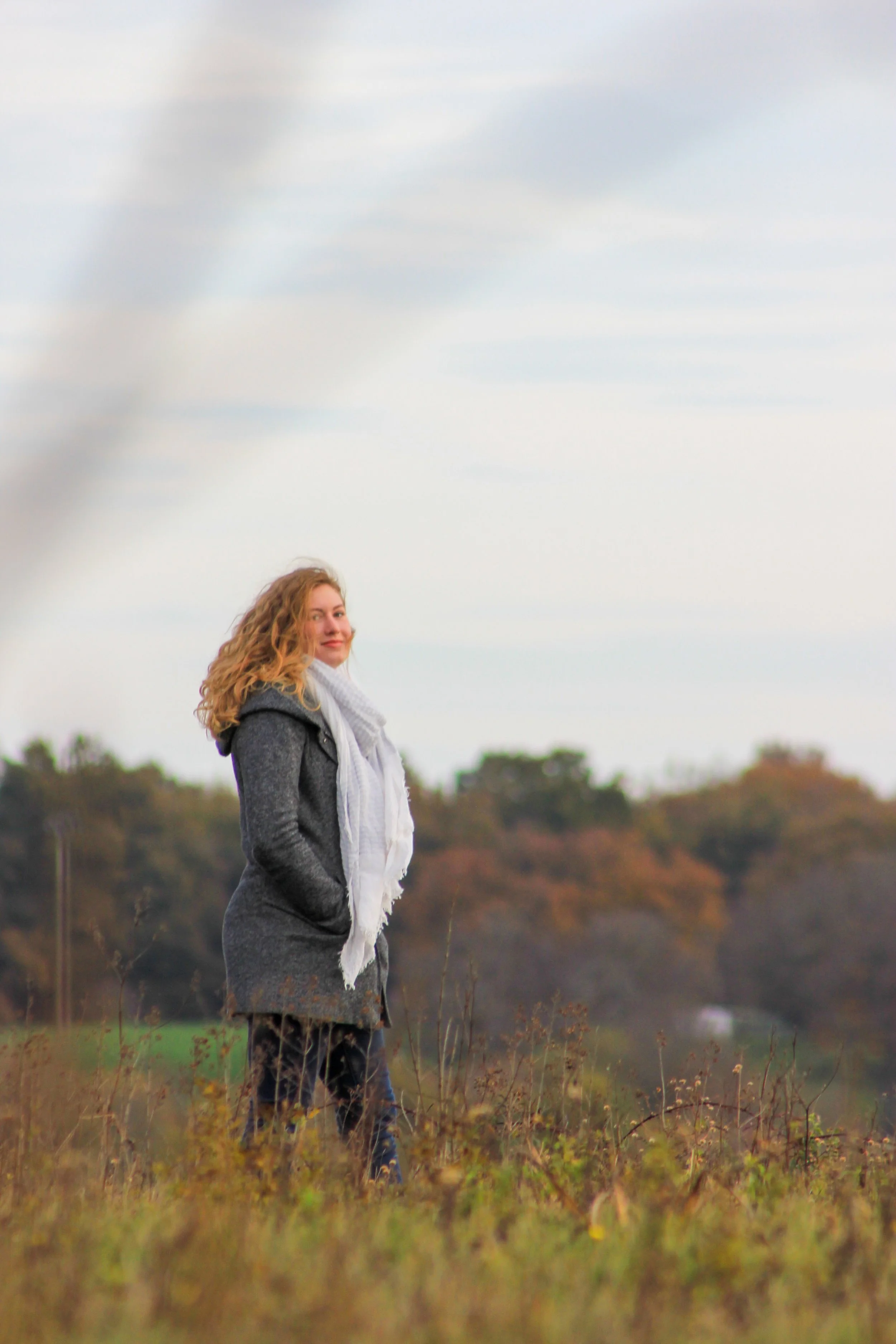 A woman with curly orange hair standing outdoors in a field, wearing a gray coat and white scarf, with her hands in her pockets, under a cloudy sky.