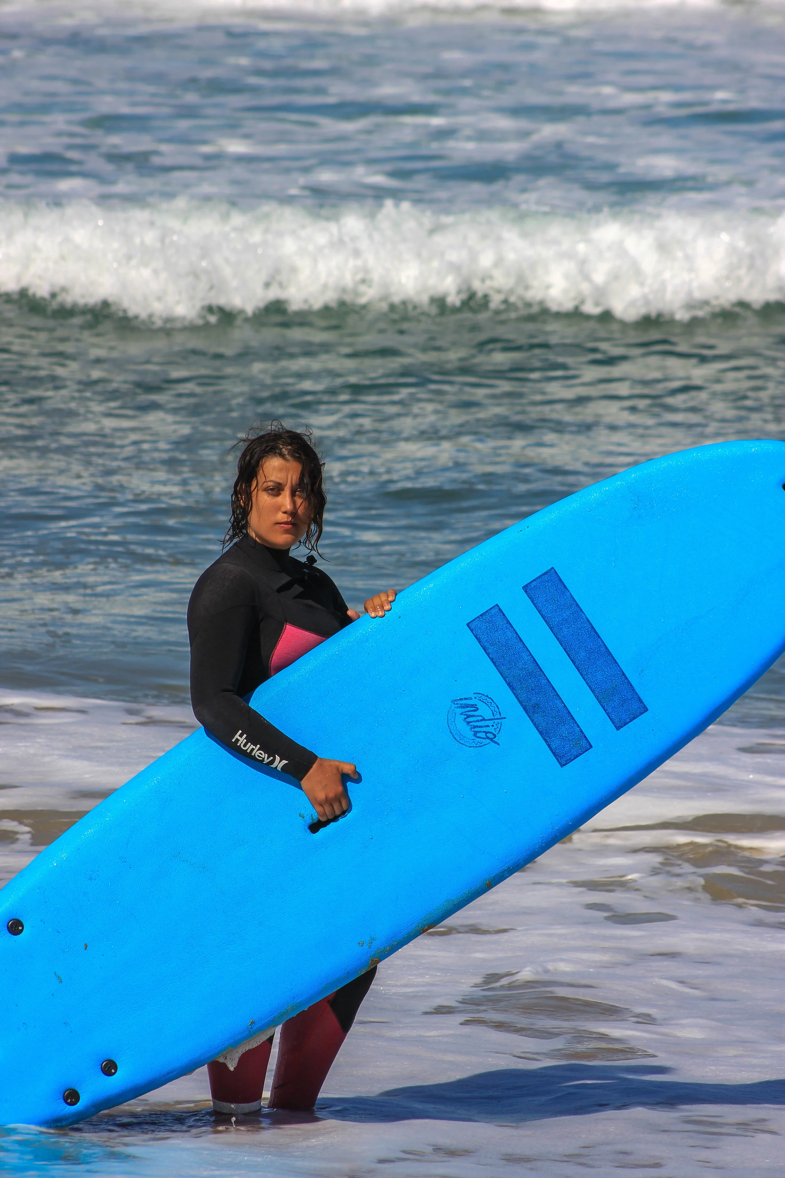 A woman holding a blue surfboard standing in shallow water at the beach with waves in the background.