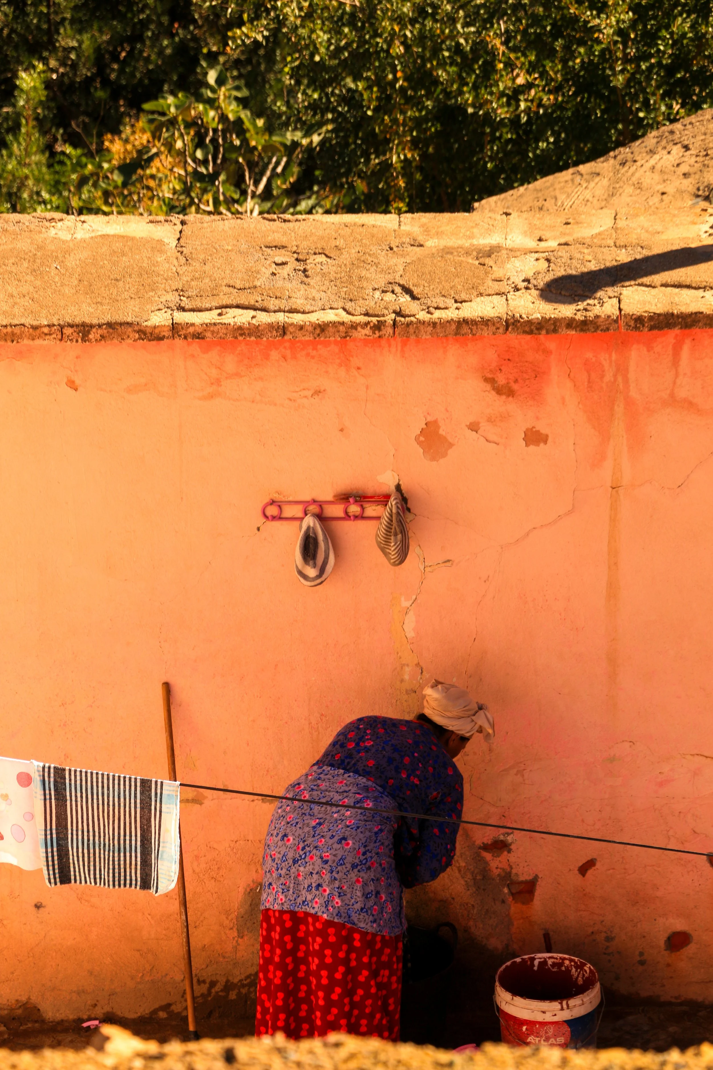 A woman wearing a headscarf and a floral dress is working on a pink wall with cracks, with some shoes hanging on a pink hook and laundry hanging on a clothesline.