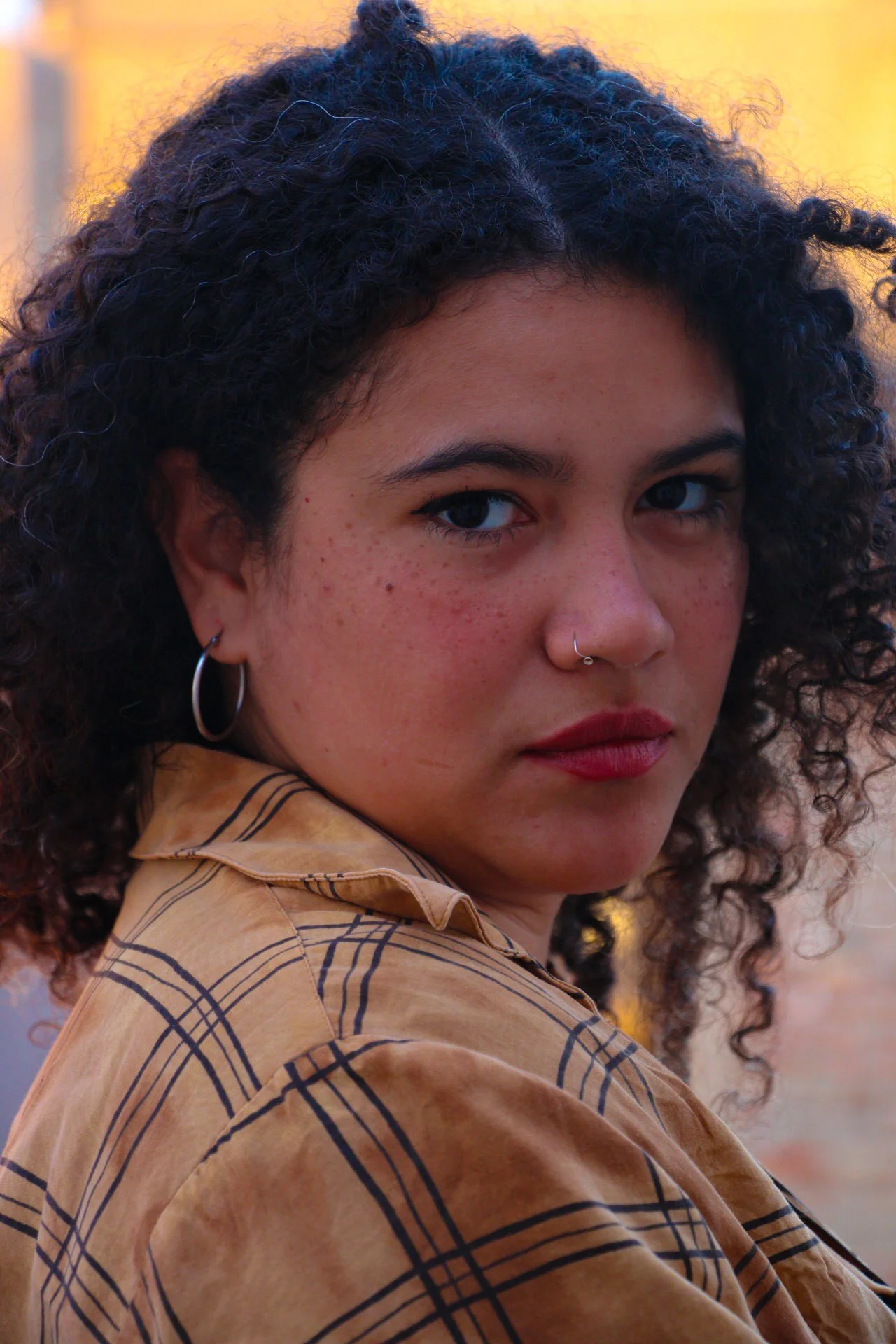 A woman with curly dark hair, wearing hoop earrings and a nose ring, looking at the camera with a serious expression. She has freckles and is dressed in a tan shirt with a black grid pattern.