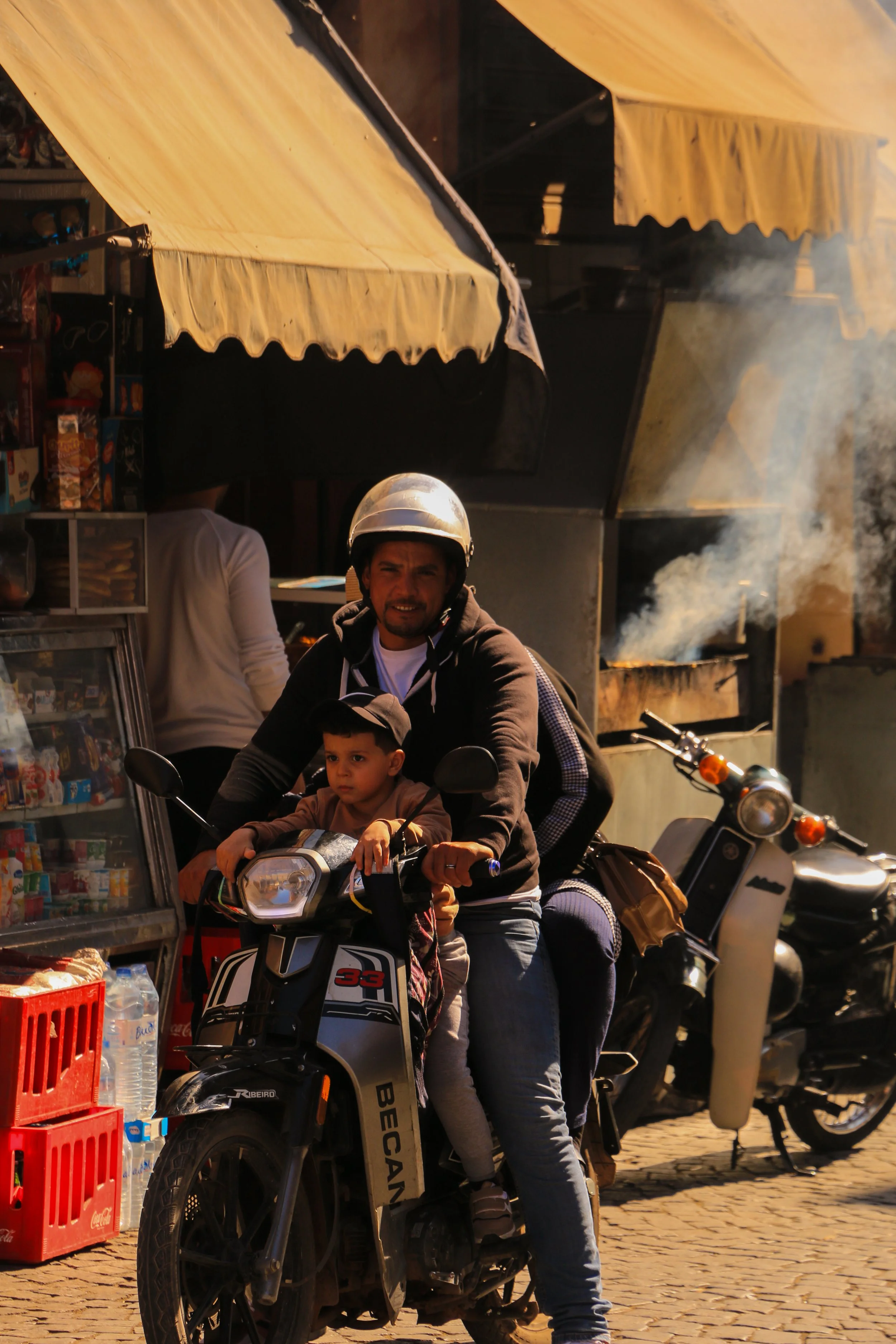 A man and a child riding a black and white scooter on a street market. The man wears a helmet and the child sits in front of him. There are market stalls with snacks and bottled water nearby, and a motorcycle parked in the background.