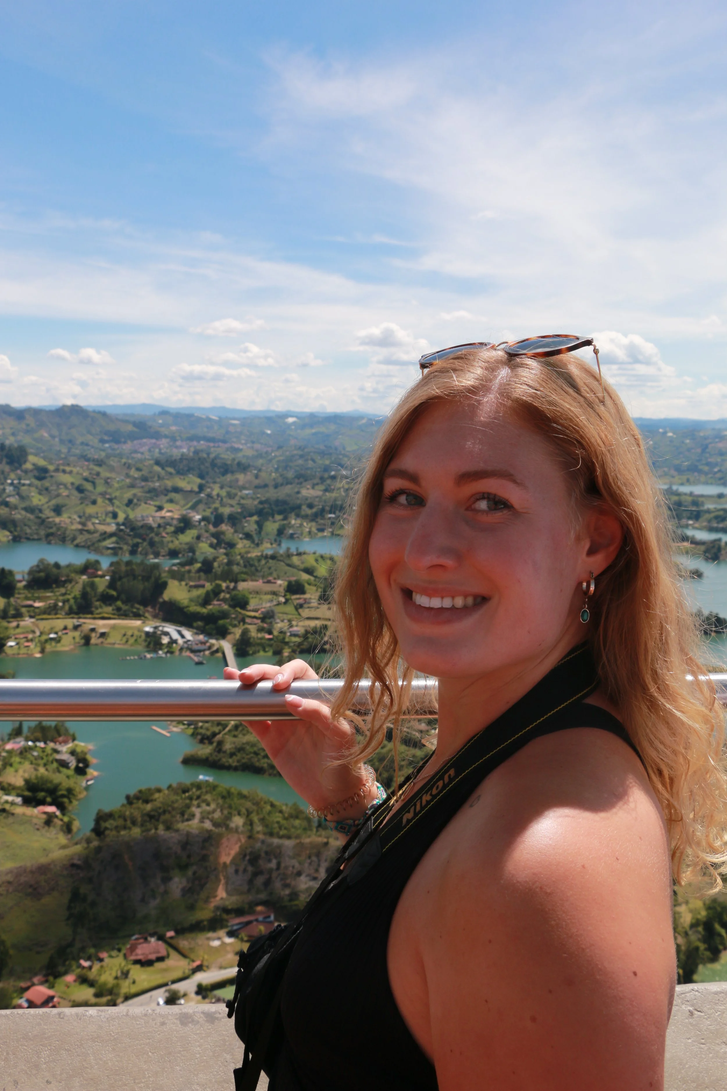 Smiling young woman with sunglasses on her head and a camera around her neck, overlooking a scenic landscape of lakes, hills, and houses from a high vantage point during daytime.