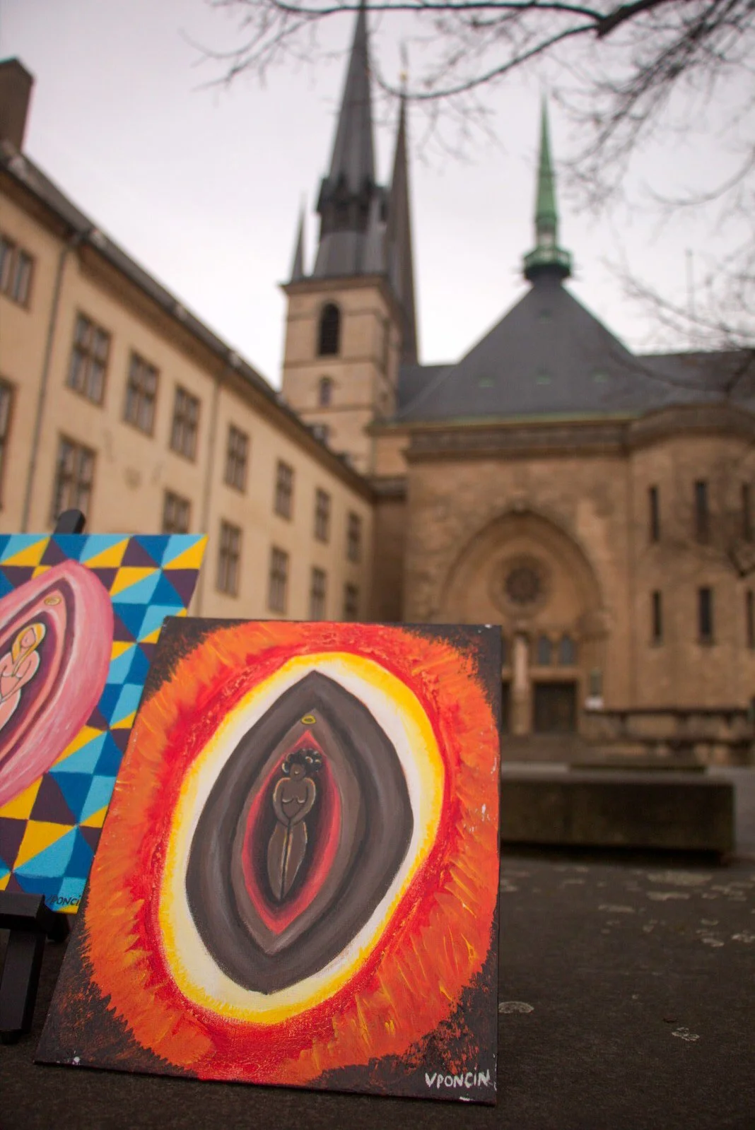 Paintings displayed outdoors in front of a historic church with two tall spires, one with a green top, and a stone facade, on a cloudy day with bare tree branches overhead.