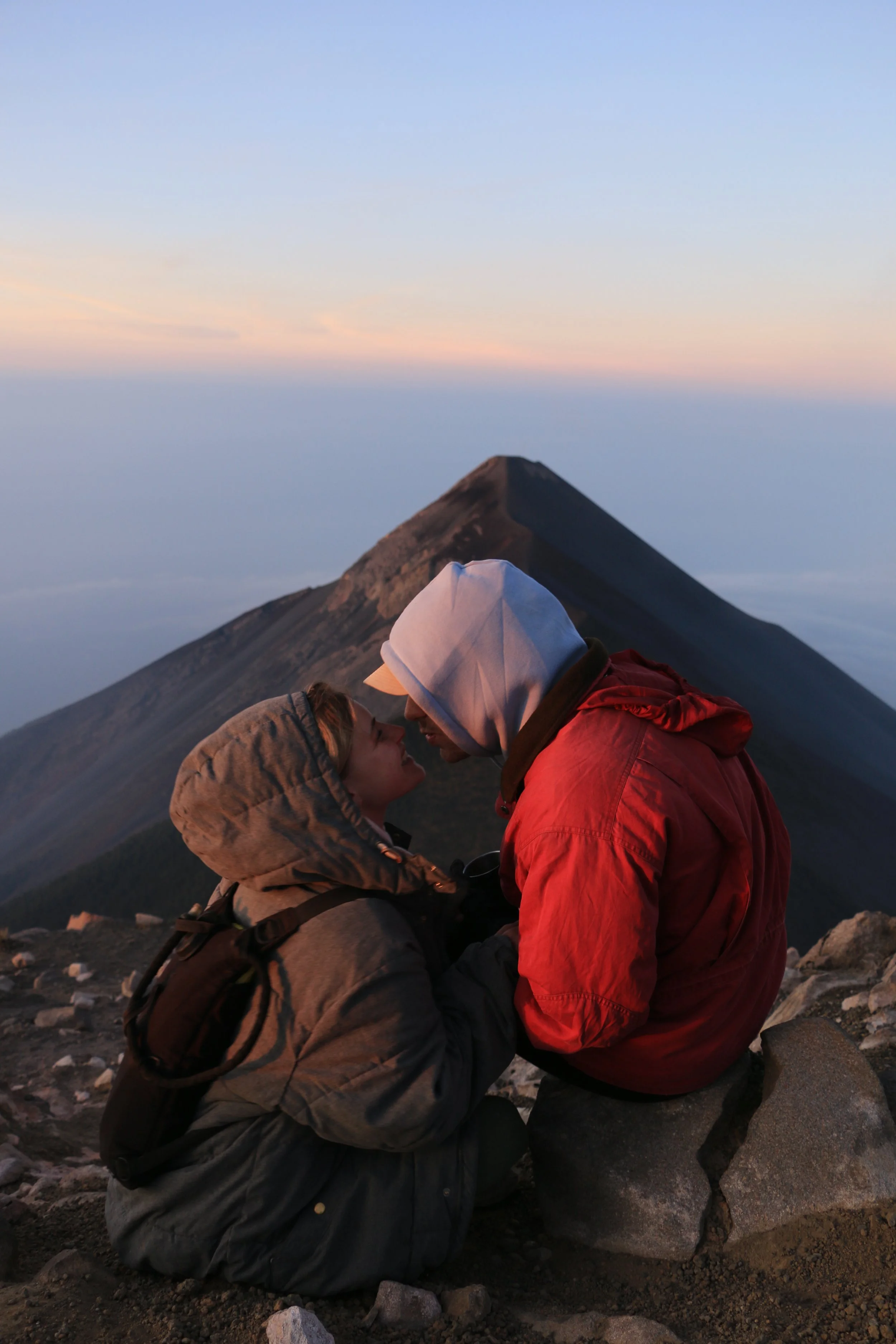 A couple sitting on rocks, facing each other with foreheads touching, at a mountain summit during sunrise or sunset with a mountain peak in the background.