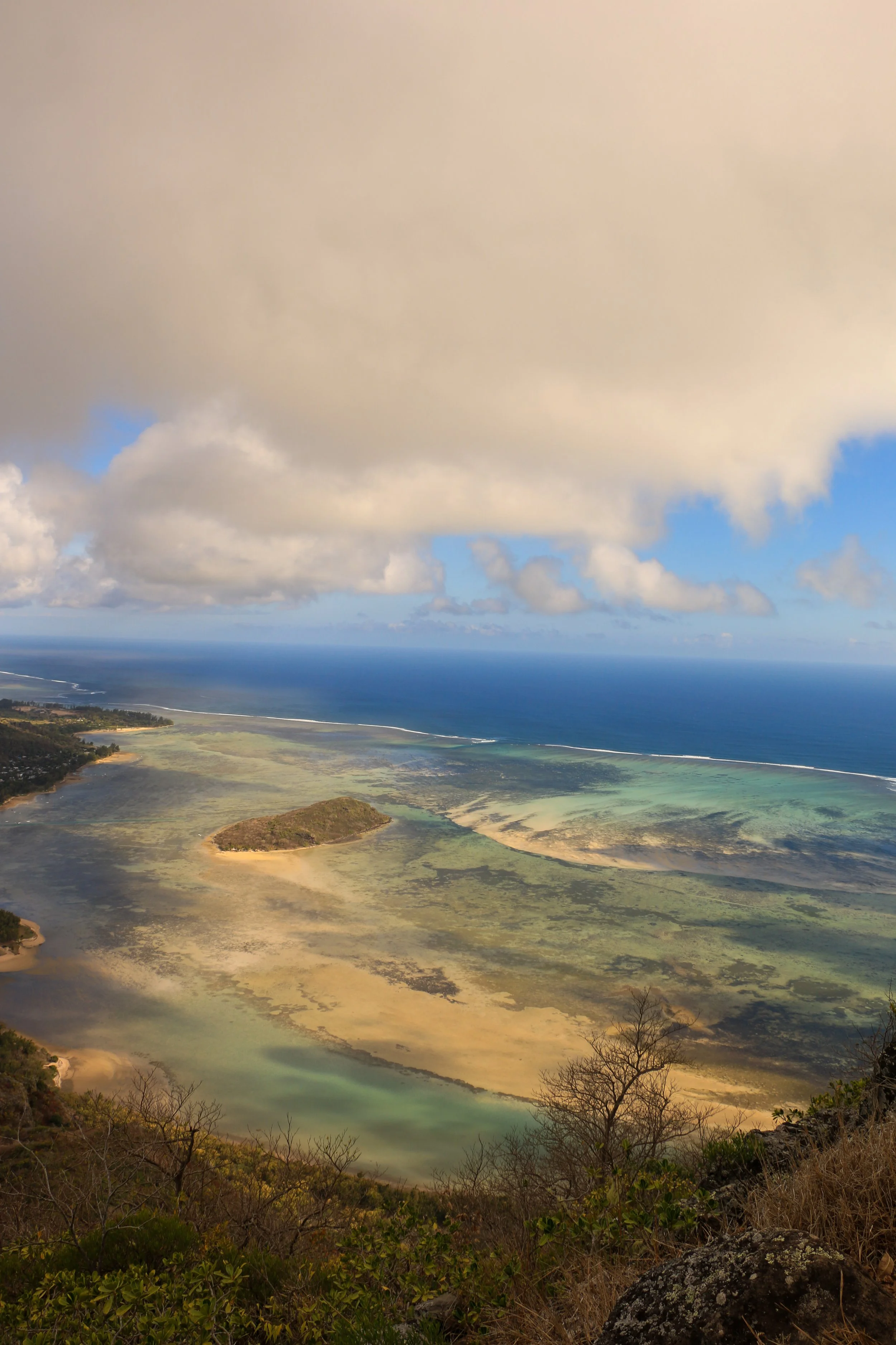Aerial view of a coastal landscape featuring a small island, coral reef, and shallow waters with clouds overhead and the ocean in the background.
