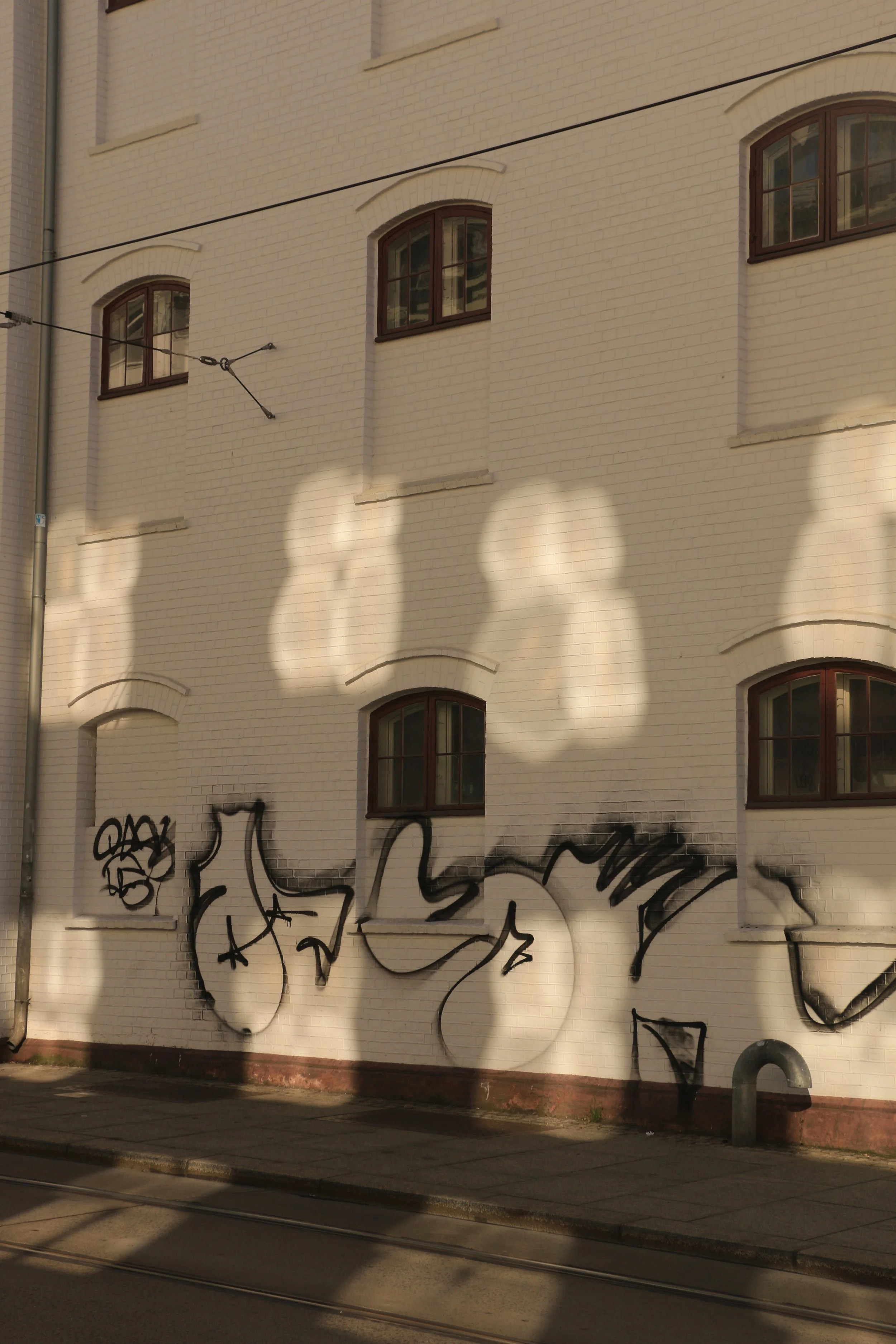 Graffiti artwork on a white brick building wall, with shadow patterns cast by sunlight on the wall and tram tracks in the foreground.