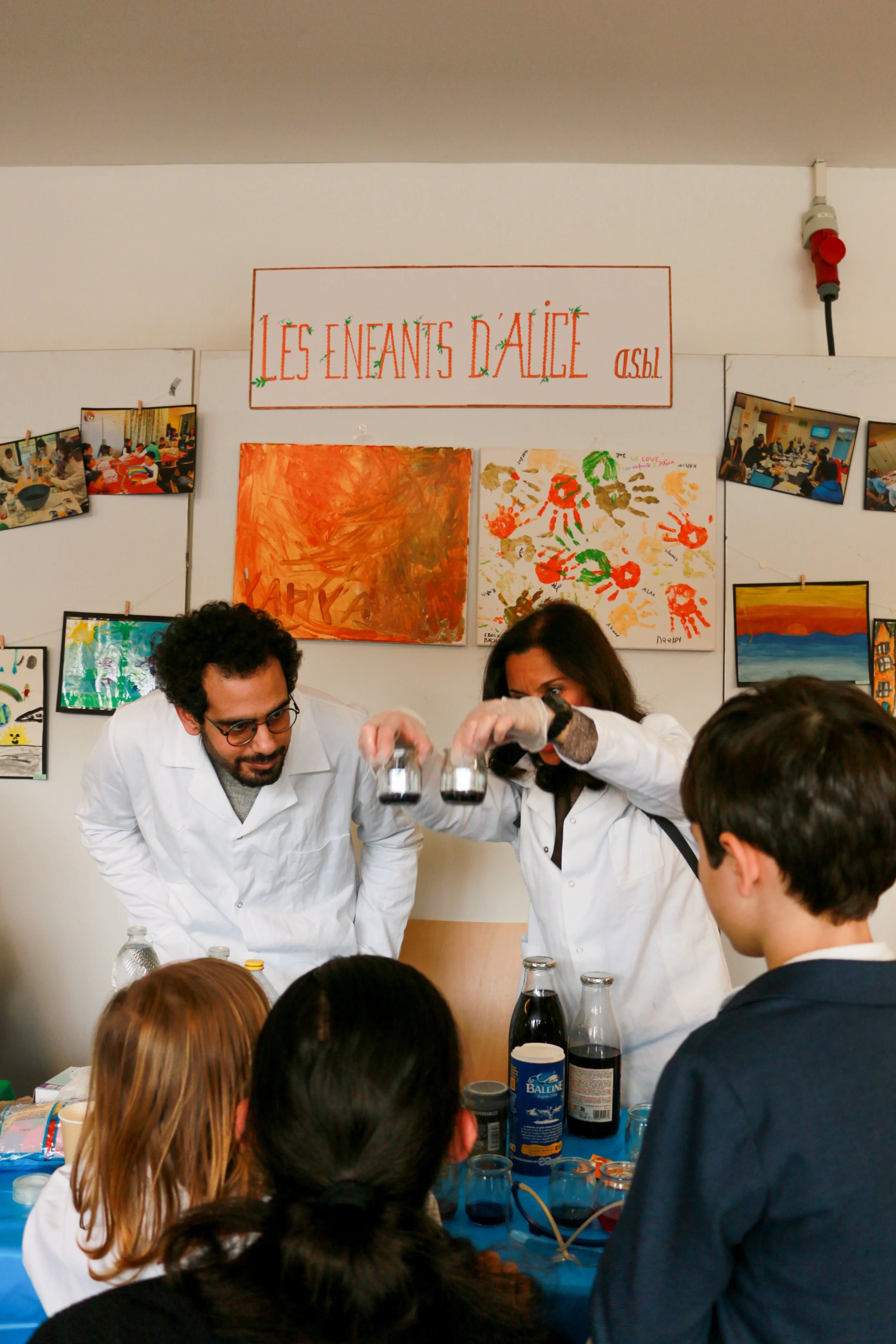 Two scientists in white lab coats conducting an experiment for children at a science event, with children watching and colorful children's artwork in the background.