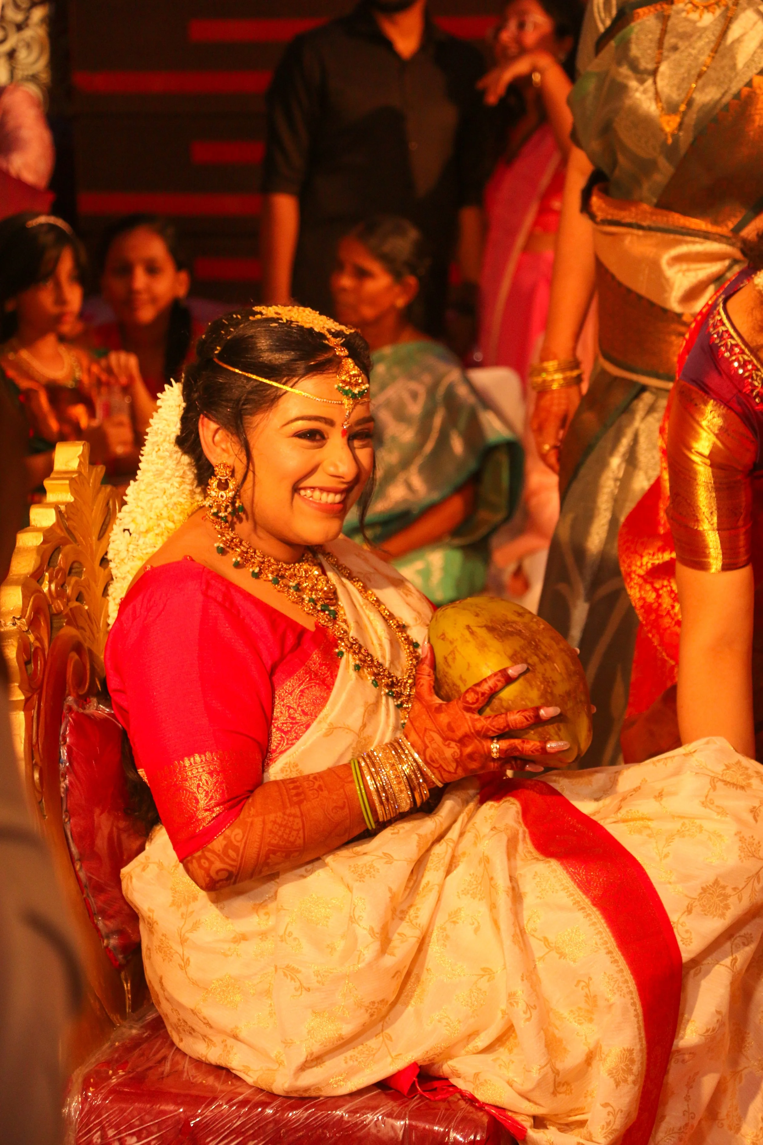A woman dressed in traditional Indian attire, wearing jewelry and floral hair decoration, sits on a throne-like chair, smiling, holding a coconut, with other people in traditional clothing around her during a festive event.