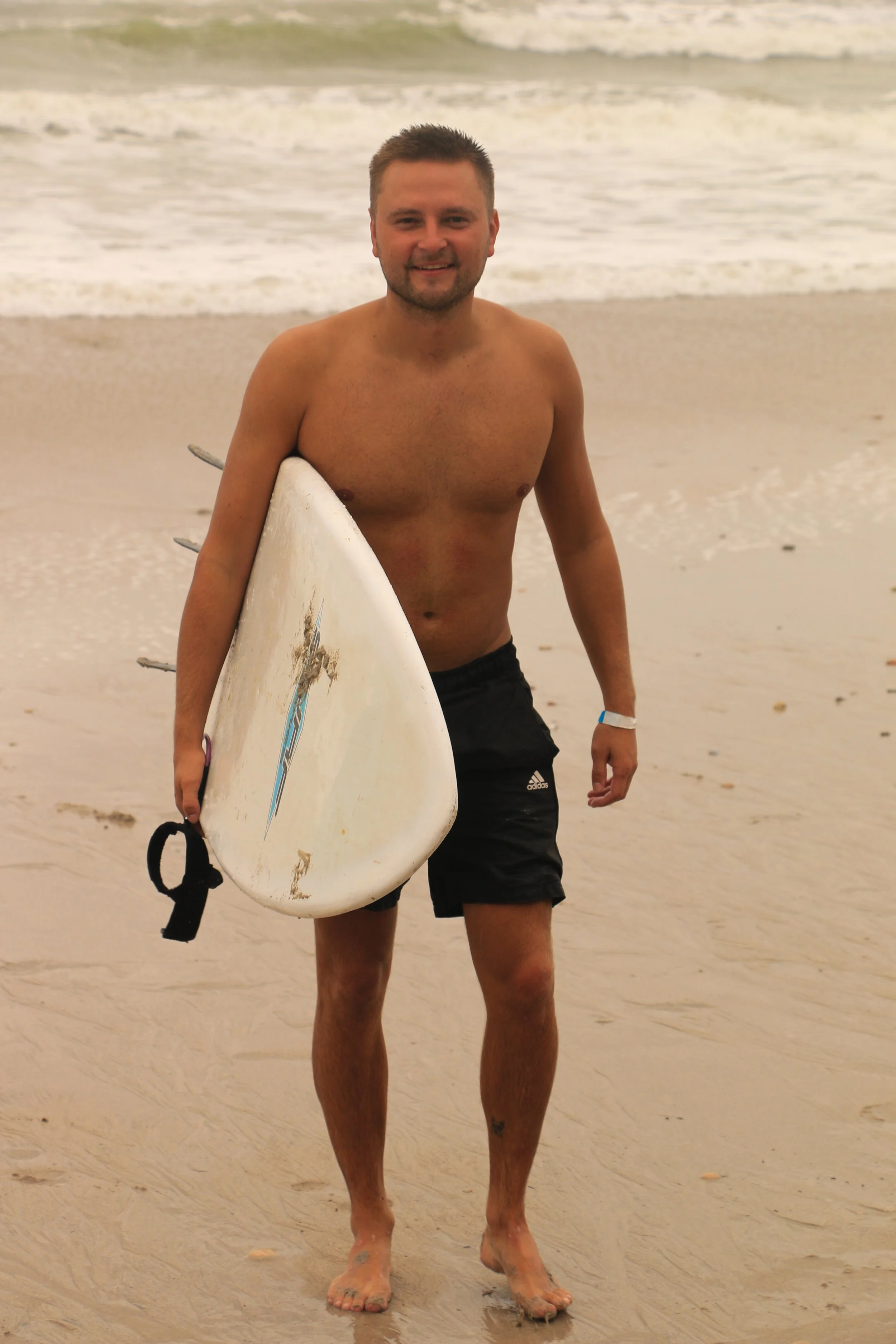 Man standing on a sandy beach holding a surfboard and smiling, with waves in the background.