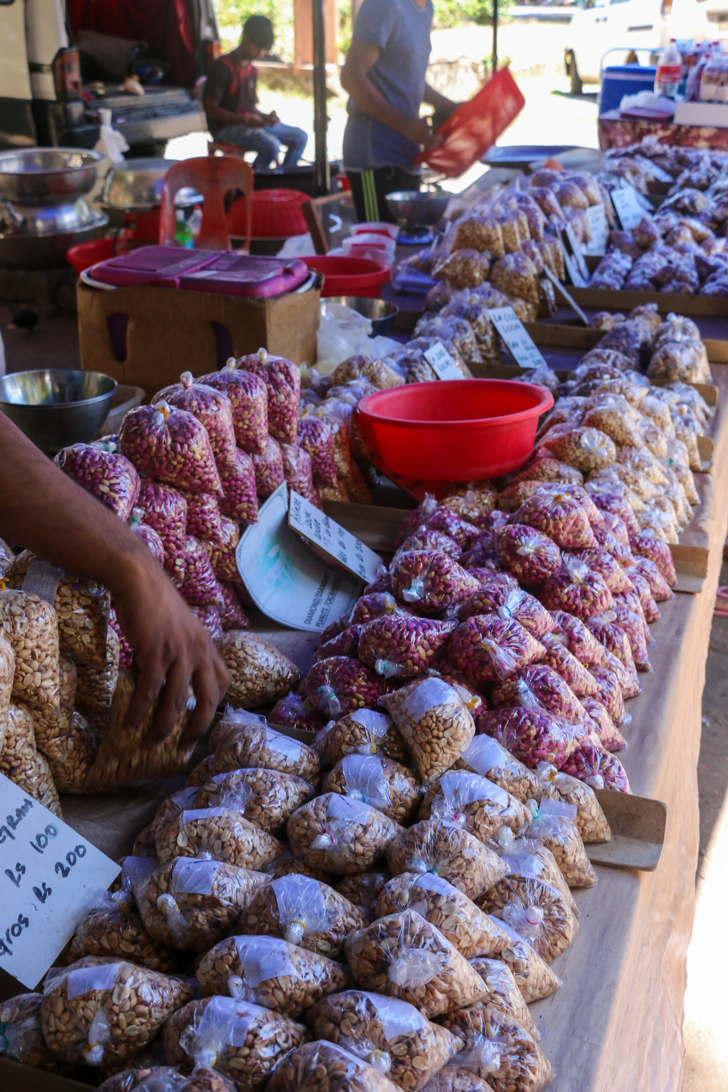 A market stall displaying various types of packaged nuts and dried fruits in plastic bags, with handwritten price signs. In the background, there are two people, one of whom is sitting and the other is standing and holding a red basket, under a canopy outdoors.