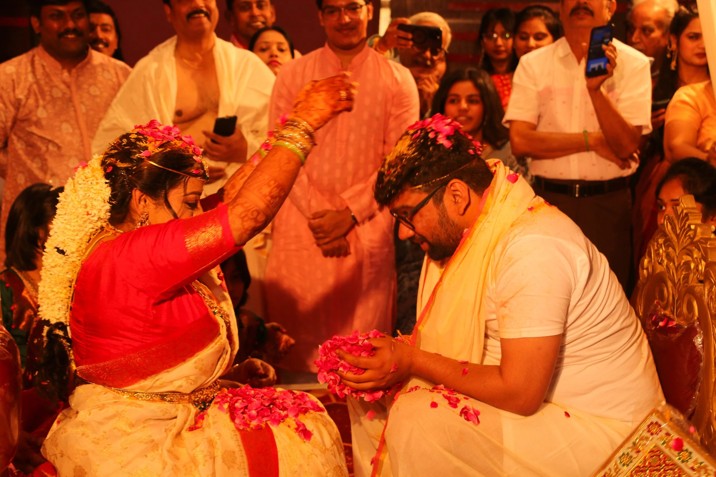 Indian bride and groom participating in a wedding ceremony, with the bride applying a floral decoration on the groom's head while both are seated, surrounded by family and friends in traditional attire.