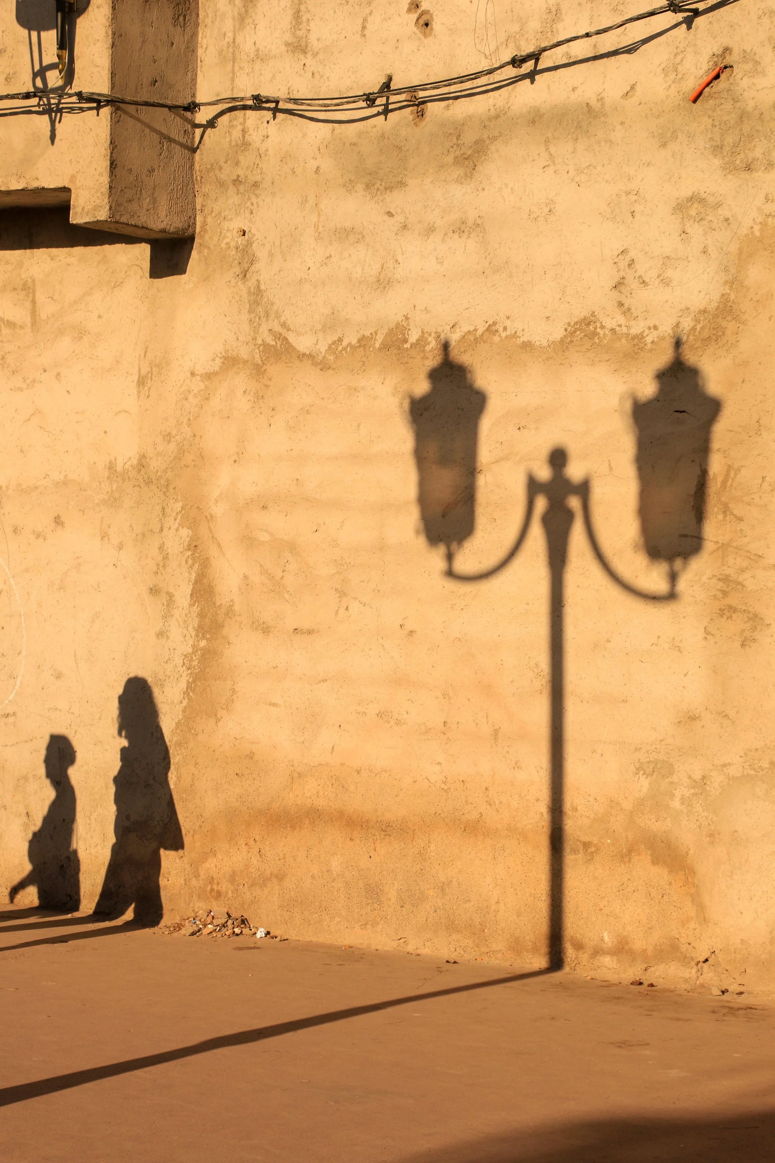 Shadows of two children and a vintage street lamp on a yellow wall, with the lamp's shadow in the center.