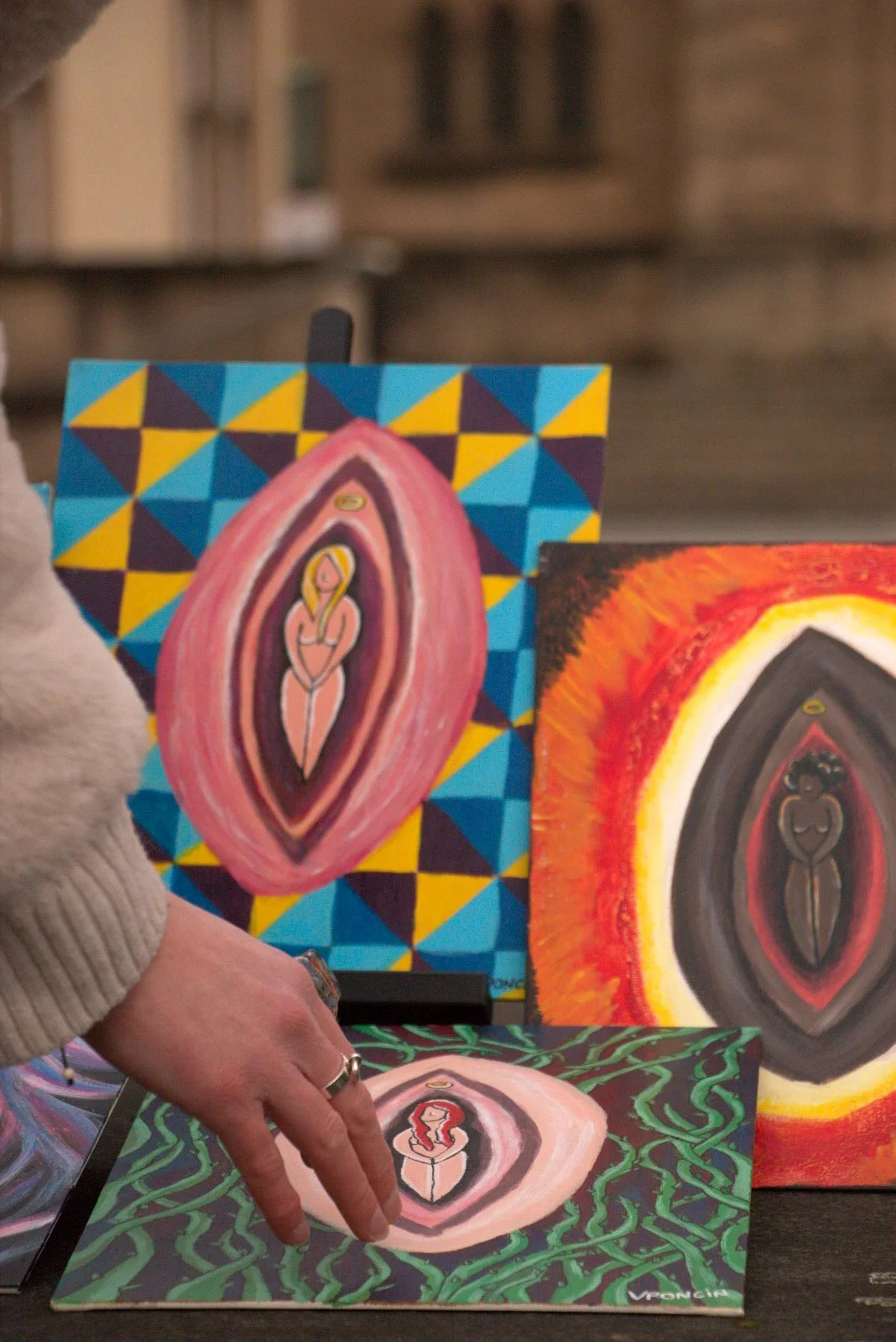 Three paintings featuring abstract female figures with halos, surrounded by colorful backgrounds, displayed on tables at an indoor art event.