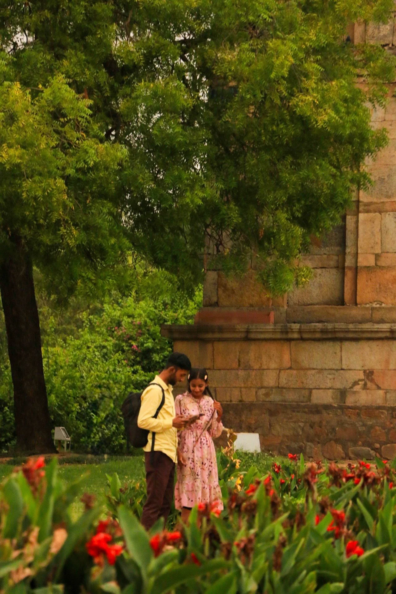 A young man and woman are standing in a garden, looking at a mobile device together. The woman is dressed in a pink traditional dress, and the man is wearing a yellow shirt and brown pants. They are surrounded by green foliage and red flowers, with a stone building in the background.