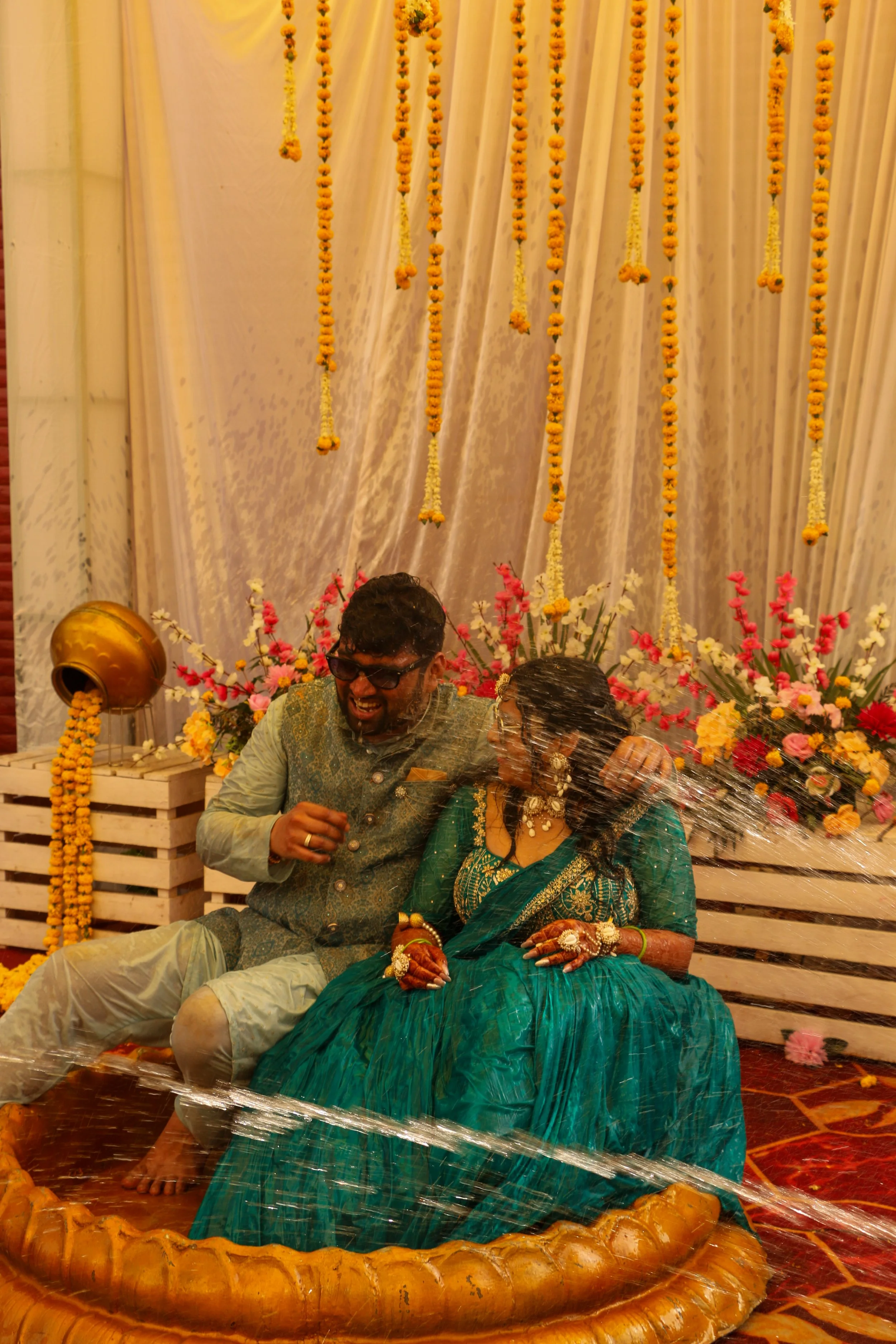 A newlywed couple seated in a traditional Indian wedding ritual, getting splashed with water, surrounded by colorful flowers and marigold decorations.