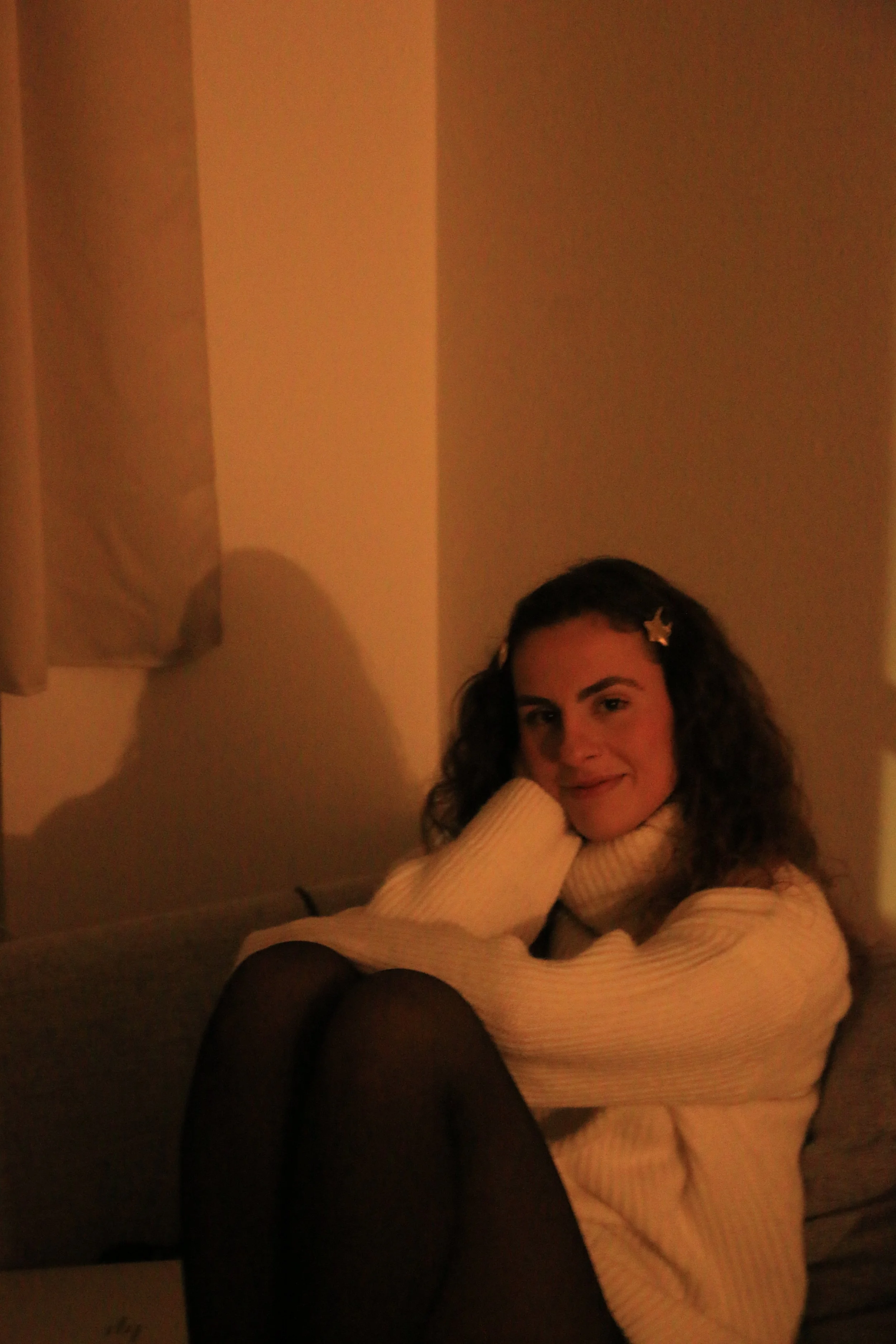 A woman with curly hair wearing a cream-colored turtleneck sweater, sitting on a sofa with her knees drawn up, smiling softly at the camera, with star-shaped hair clips in her hair, in a dimly lit room.