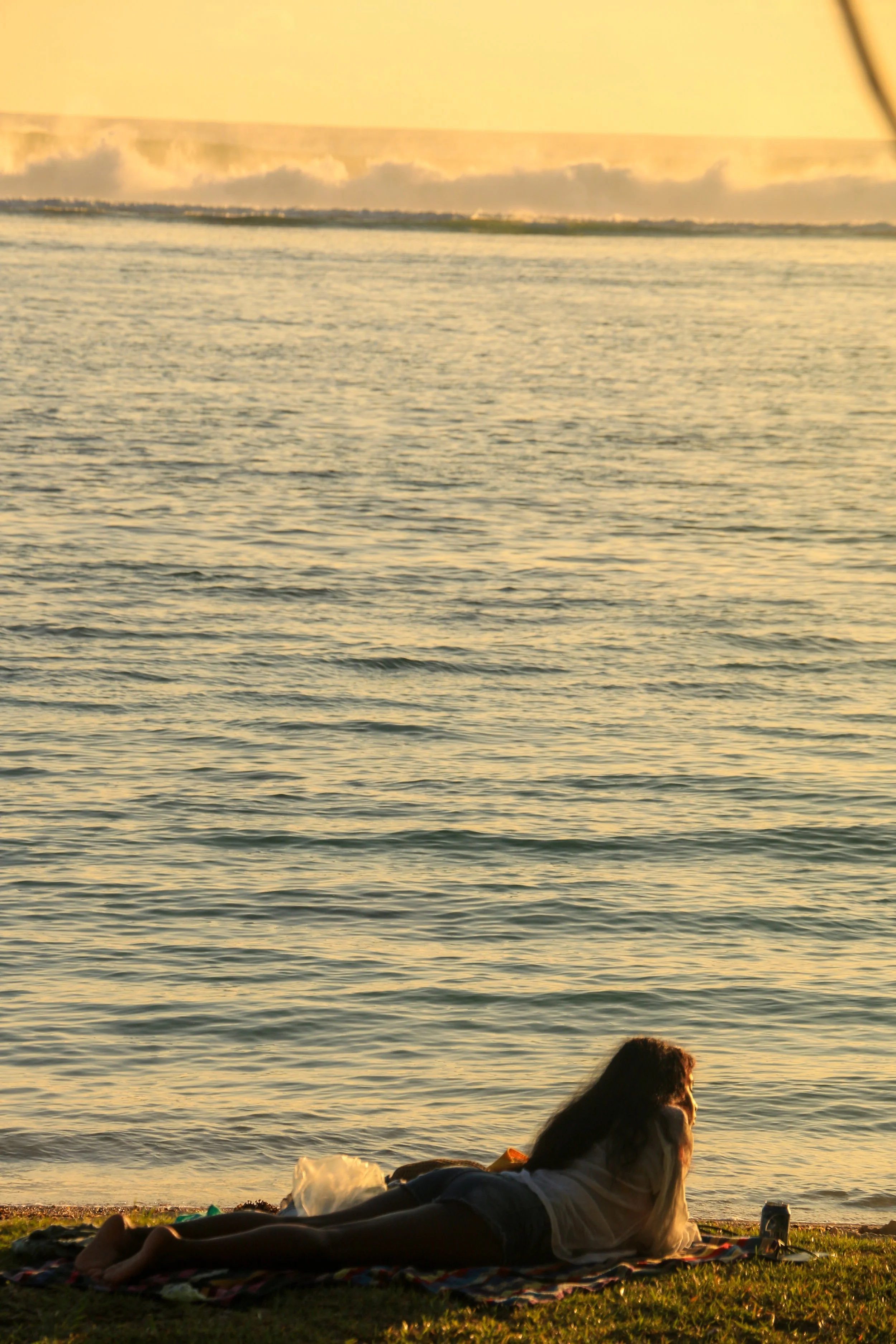 A woman lies on a blanket at the beach during sunset, facing the water with waves in the background.