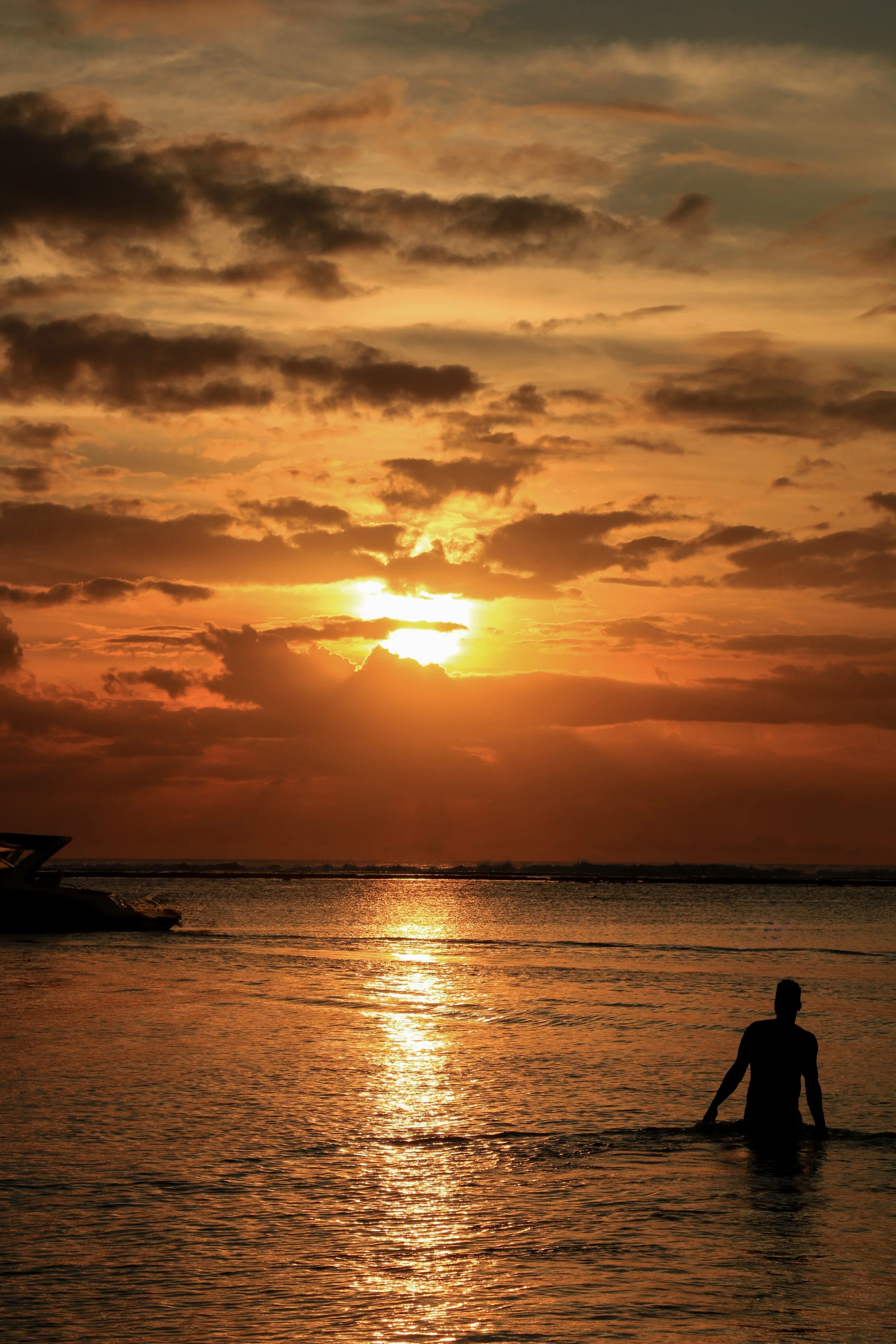 Silhouette of a person sitting in shallow water on a beach during sunset, with clouds in the sky and boats visible in the distance.