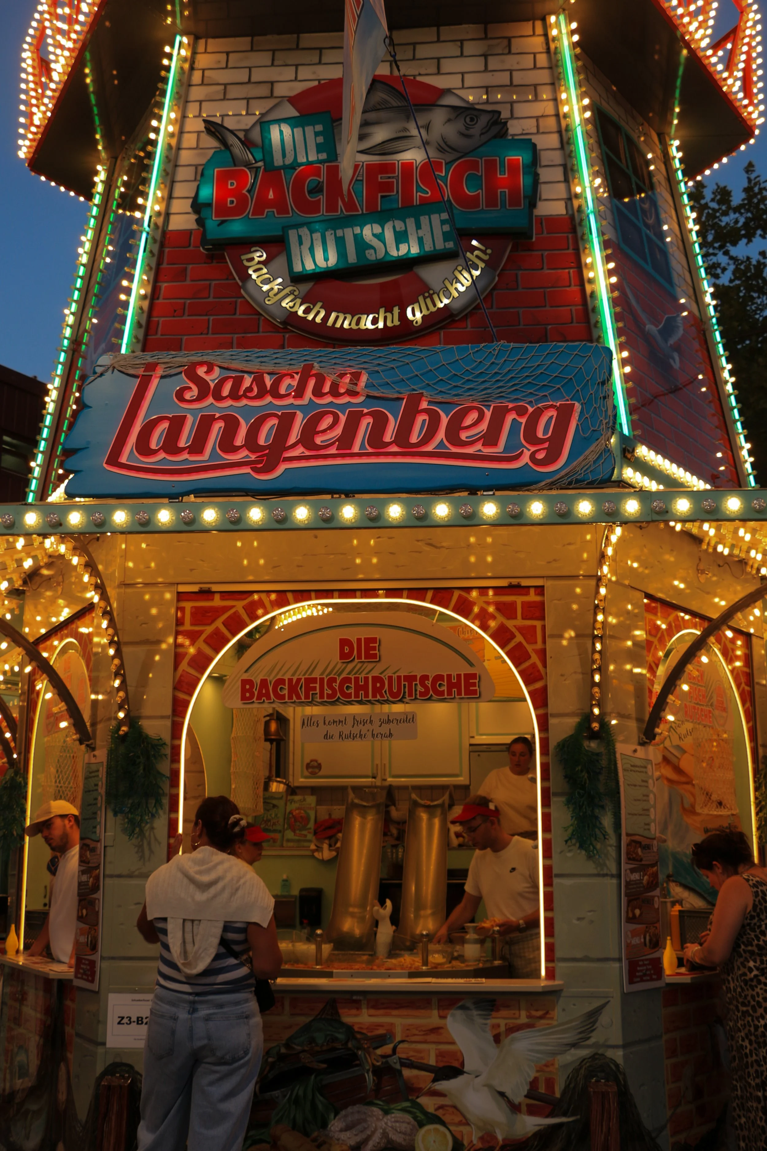 A food stall at a fair, decorated with bright lights and signs. The signs are in German and advertise "Die Backfisch Rutsche" and "Sascha Langenberg." There are people waiting in front of the stall, and a person inside preparing food.
