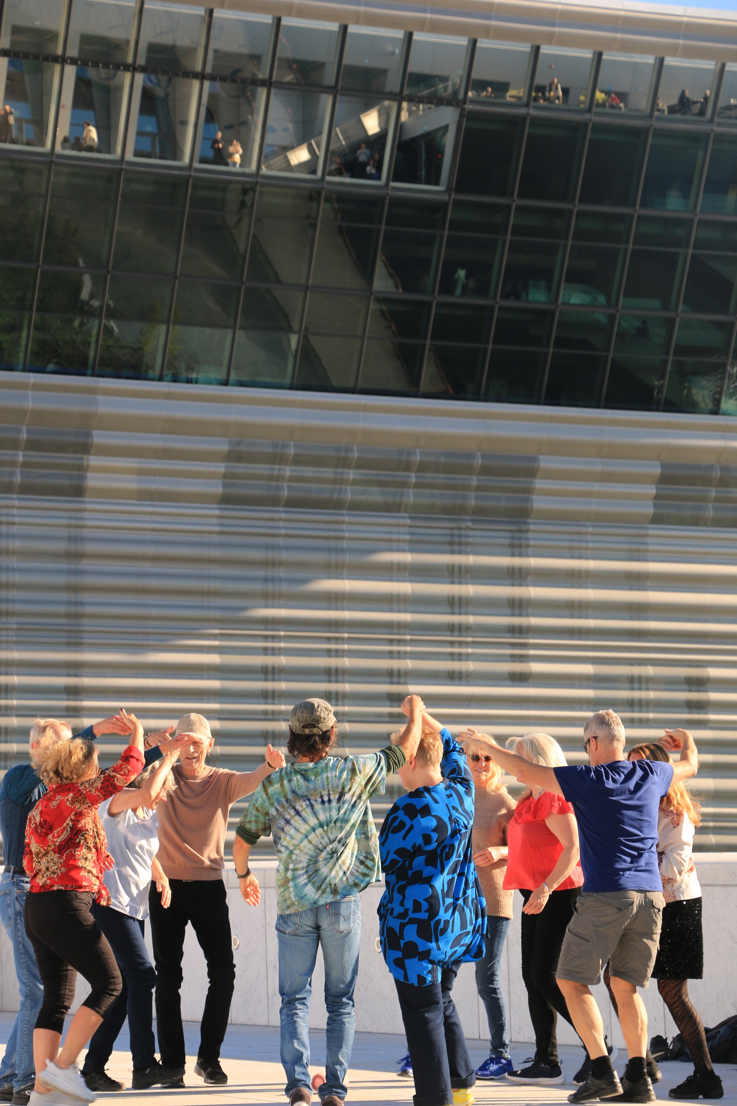 A diverse group of people dancing outdoors in front of a modern glass building with reflections.