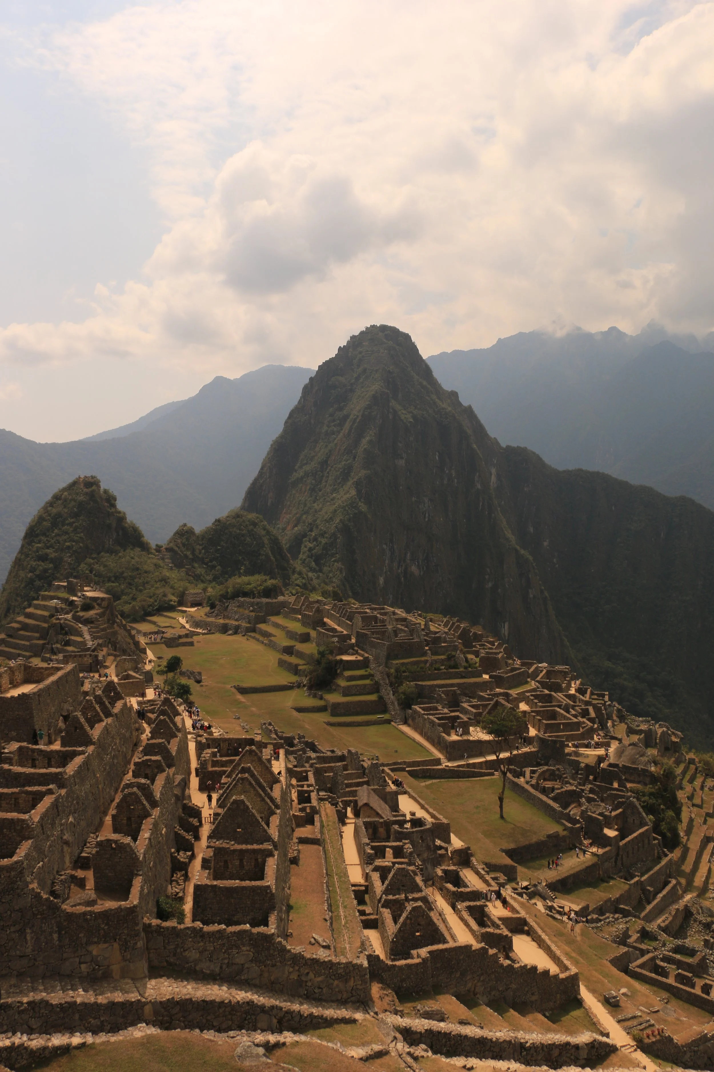Ancient ruins of Machu Picchu with tiered terraces, stone structures, and mountainous backdrop under a partly cloudy sky.