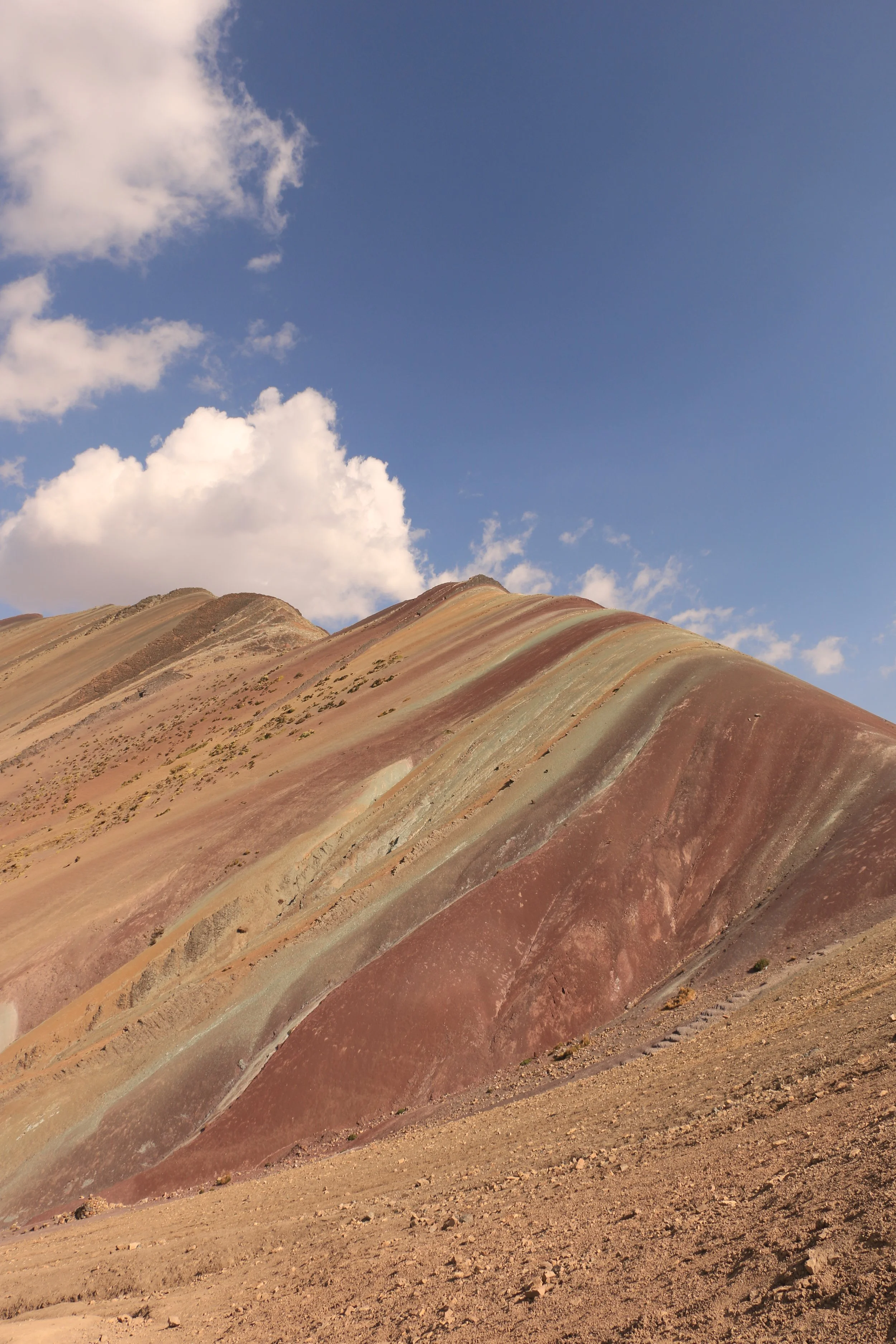 Colorful mountain with stripes of various shades of red, pink, and green under a blue sky with scattered clouds.