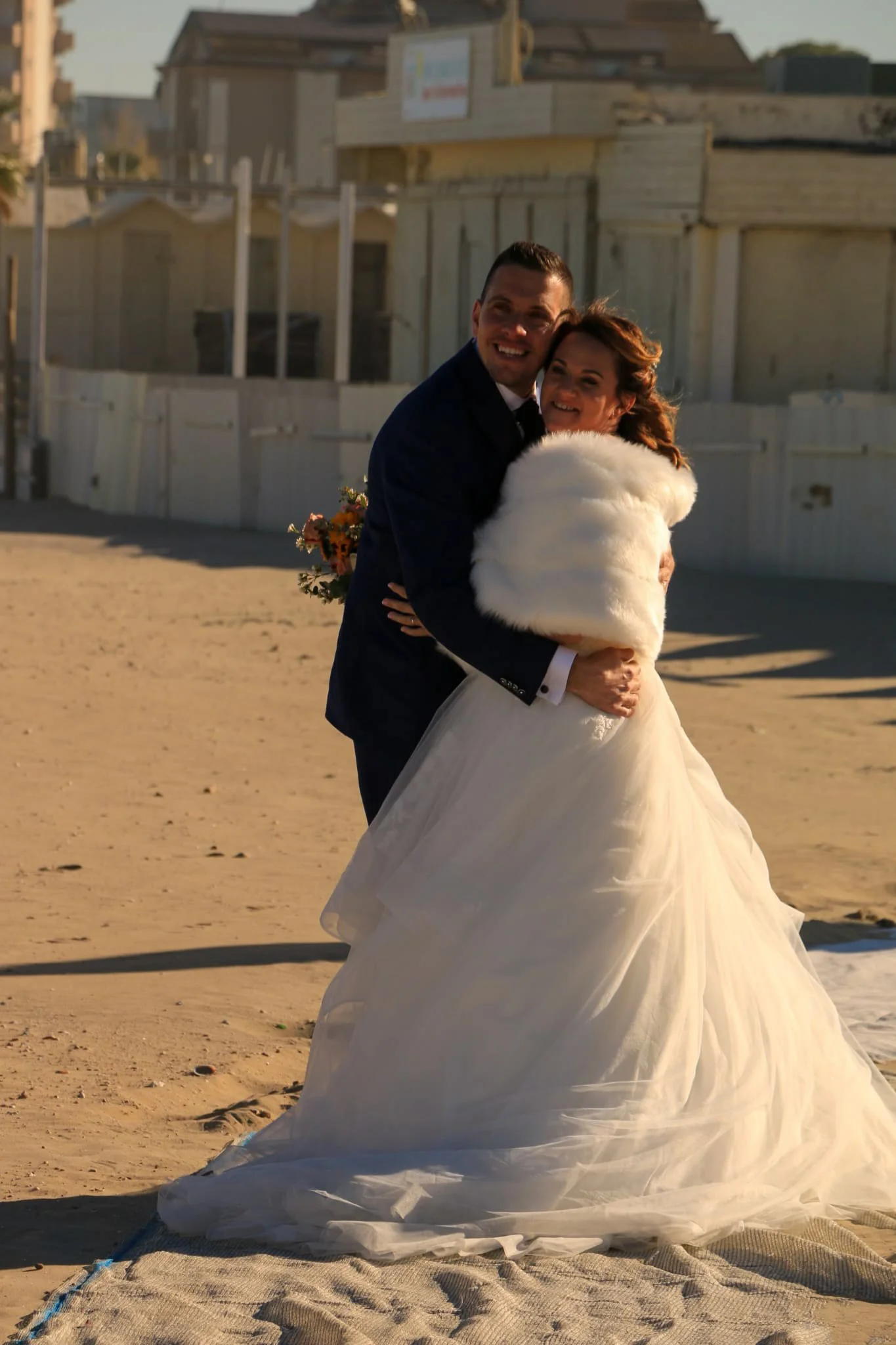 A couple in wedding attire embracing on a beach, with the bride wearing a white gown and a fur shawl, and the groom in a dark suit, in front of beach houses during sunset.