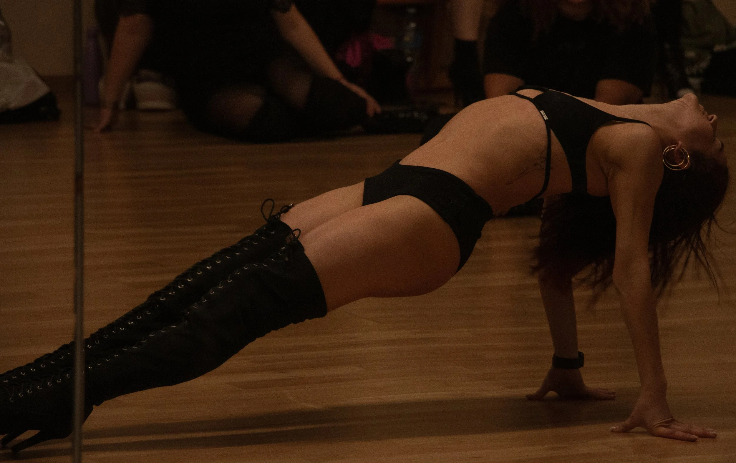 A woman with long dark hair performing a yoga pose on a wooden floor, wearing black underwear and black boots, with other people sitting in the background.