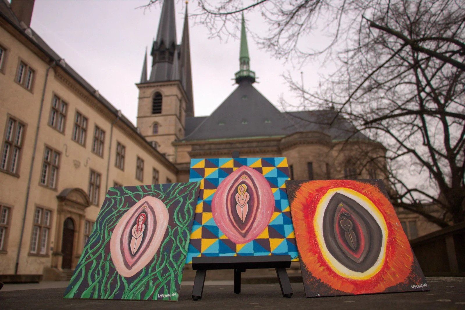 Three colorful paintings of a feminine nude figure with a halo, displayed outdoors against an old building with trees in the background.