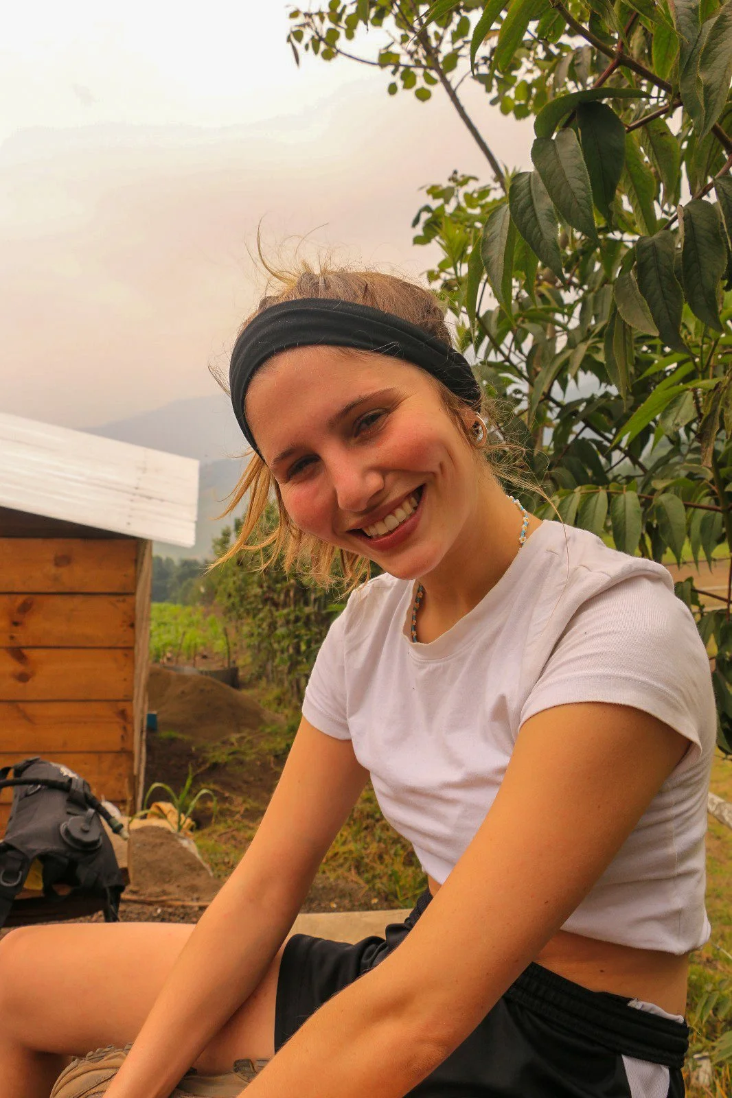 A young woman with a black headband smiling outdoors, surrounded by green foliage and a wooden structure.