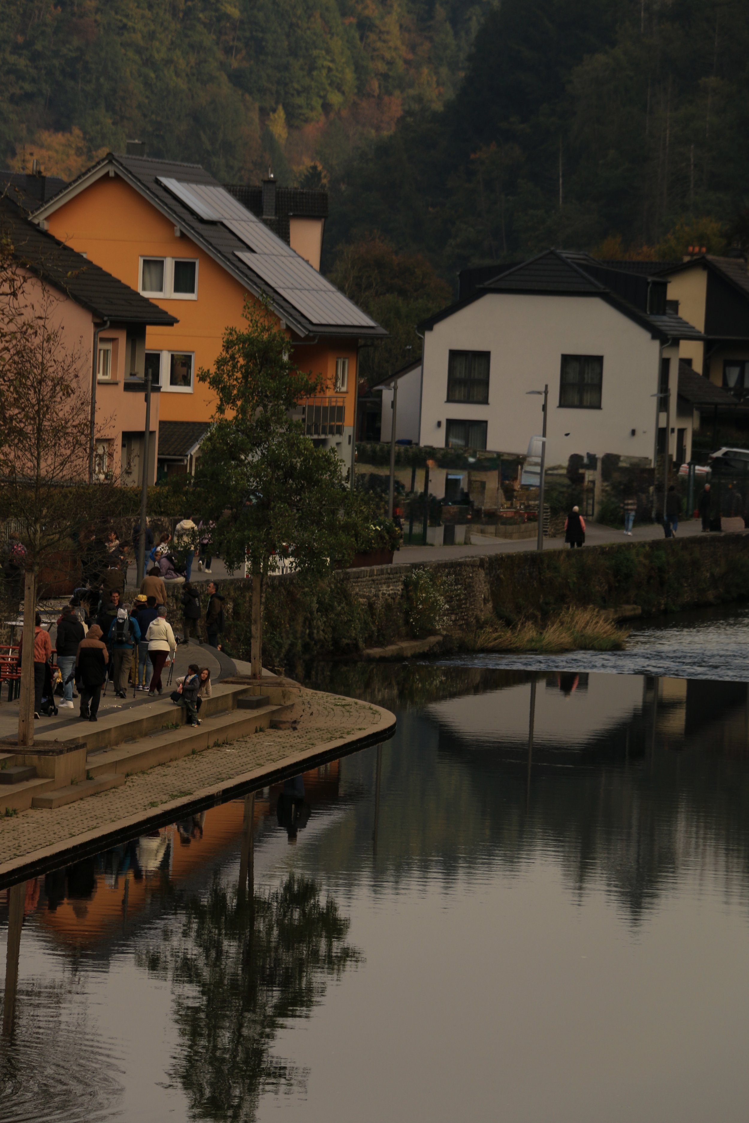 A lakeside town with colorful houses, trees on a hillside, and people walking along a paved path near the water, reflecting in the lake.