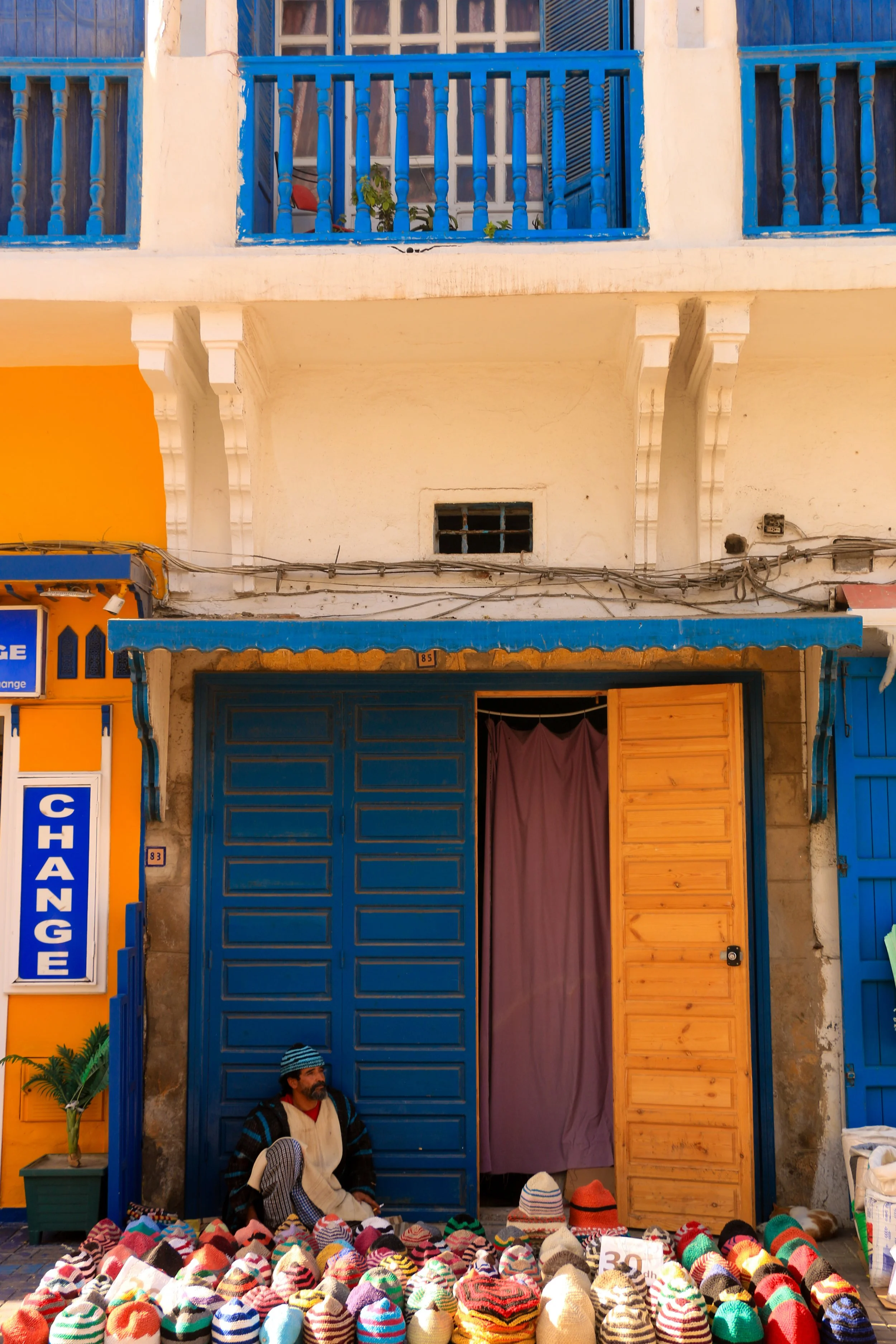 Street vendor with a beard and striped hat sitting among colorful knitted hats and scarves for sale, in front of a bright blue door with a purple curtain, on a cobblestone street.
