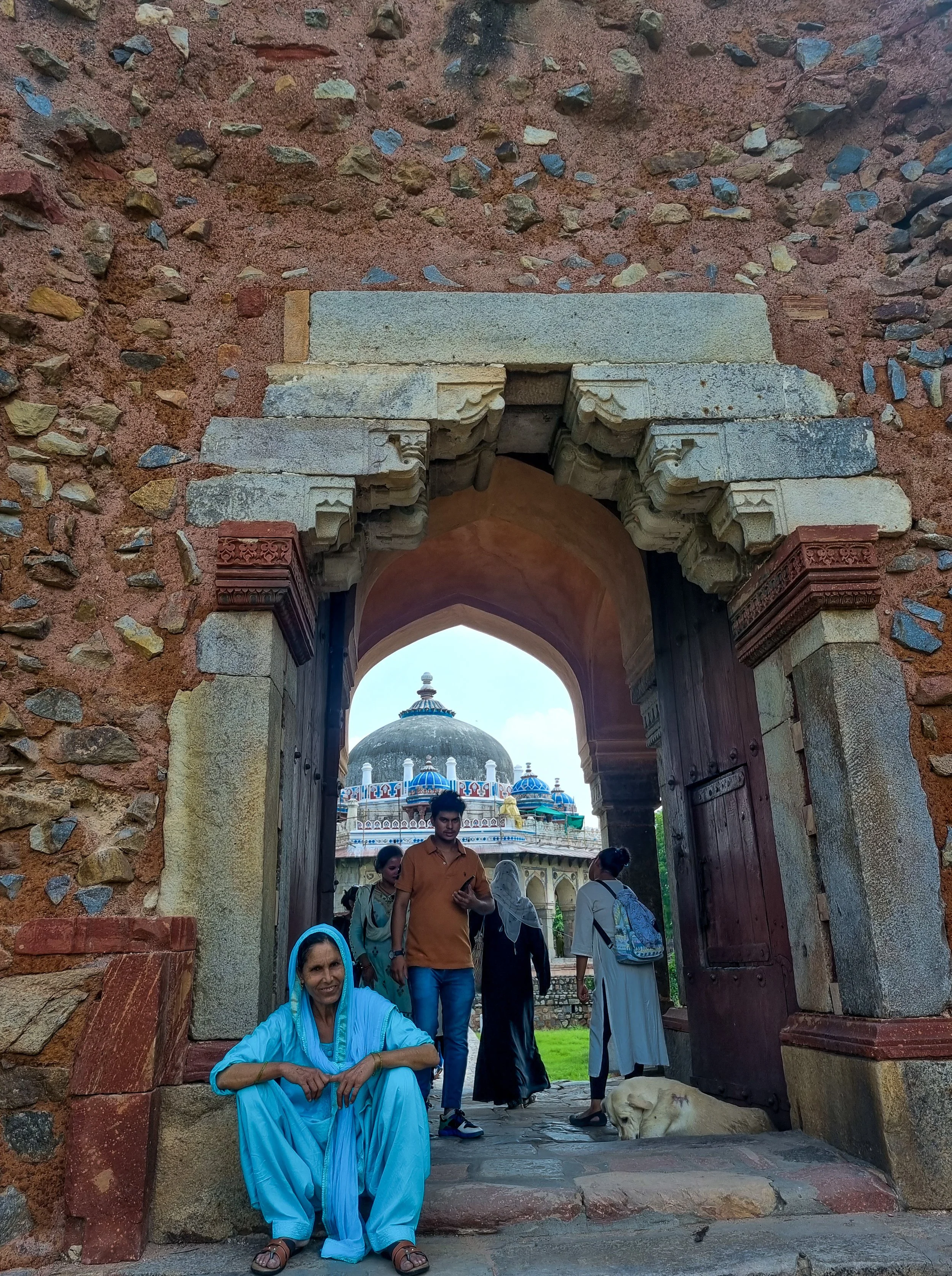 People entering a historic temple with a large dome in the background, a woman sitting in front with a blue headscarf, a dog lying on the ground inside the temple gate.