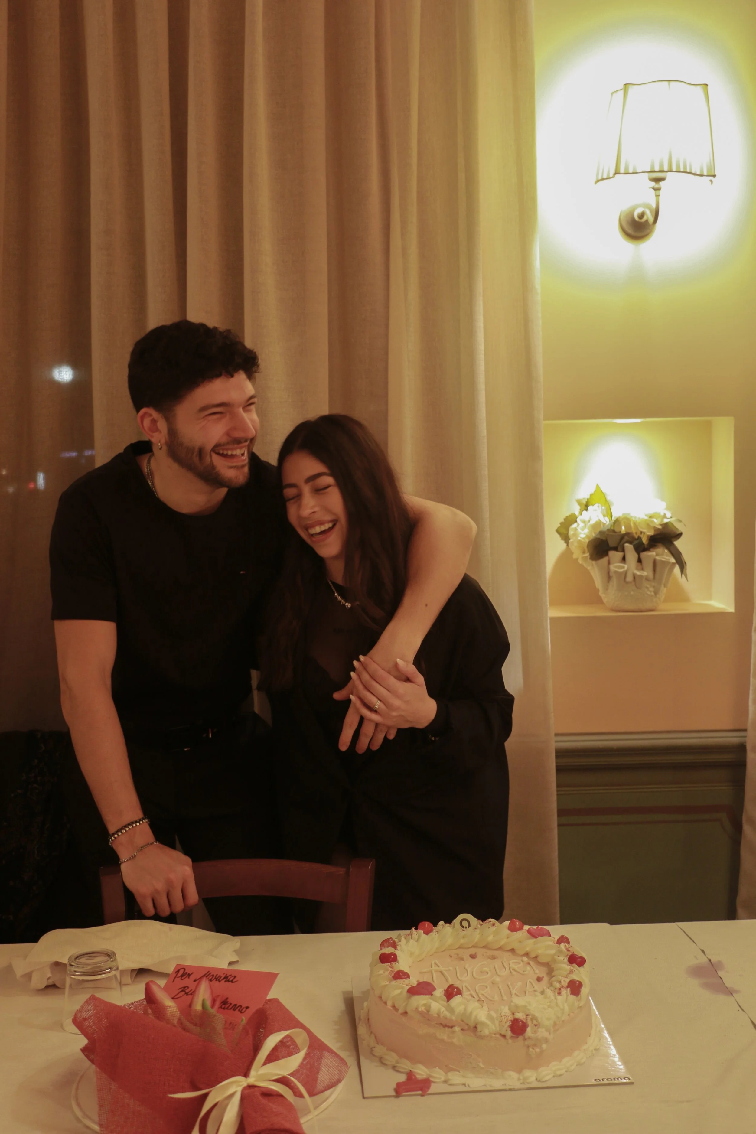 A man and woman are laughing and hugging in front of a table with a heart-shaped birthday cake and a red gift bag in a warmly lit room.