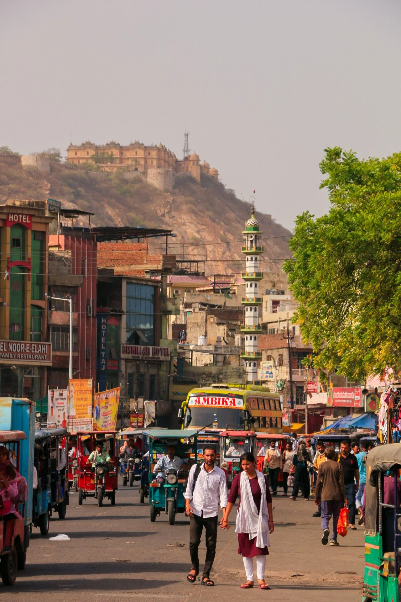 A busy street scene in an Indian city with pedestrians, auto rickshaws, and buses, with a hilltop fort and mosque minarets visible in the background.