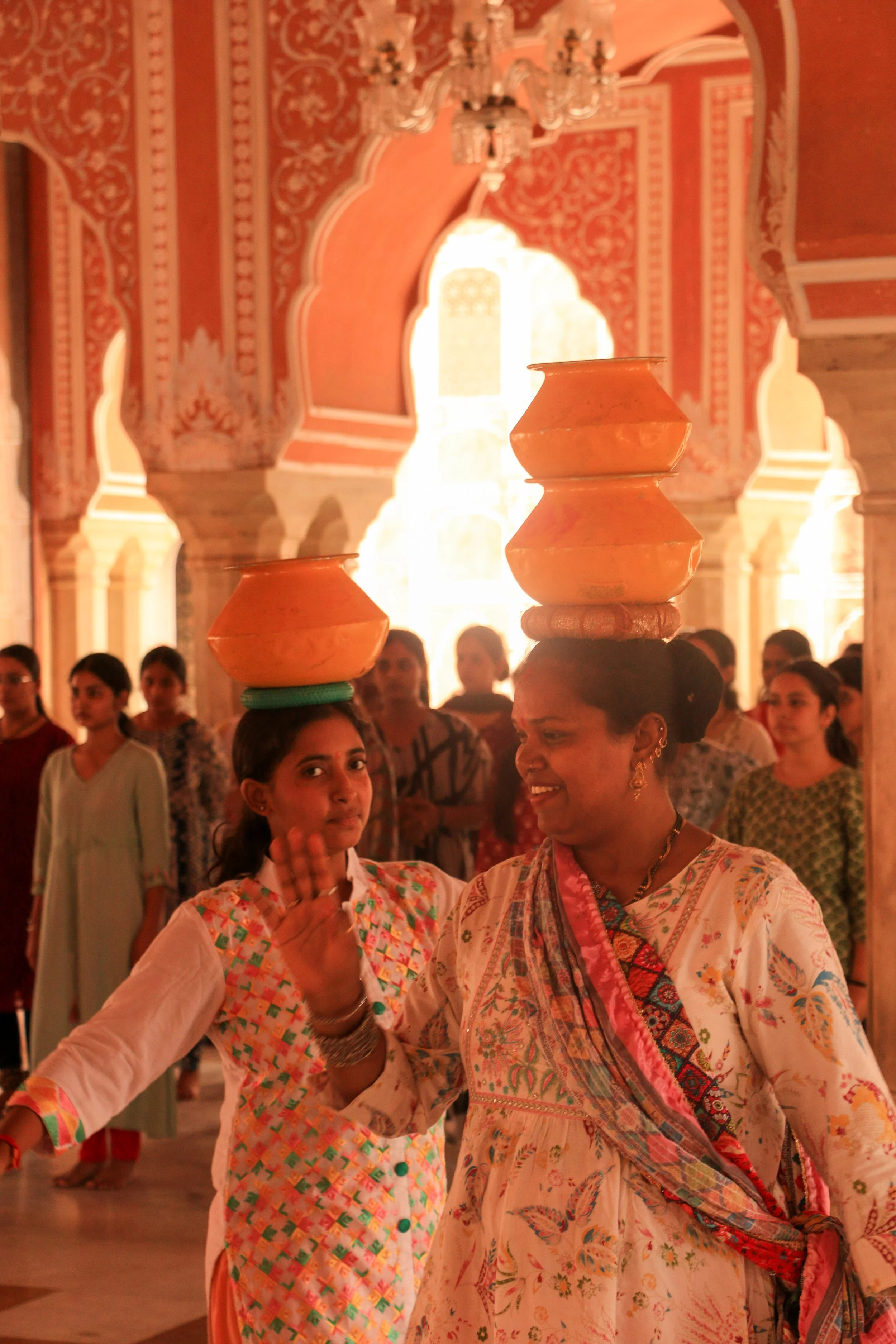 Indian women, including a woman with a pot balanced on her head and a woman raising her hand, participating in a cultural dance inside a temple or palace with intricate architecture and a chandelier.