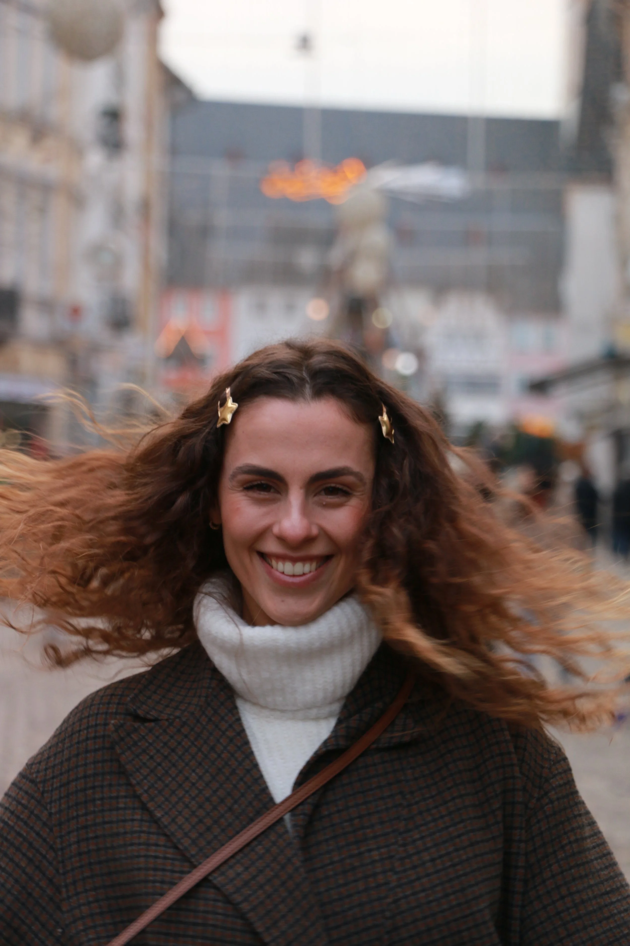 Smiling woman with curly hair blowing in the wind, wearing a white turtleneck and brown plaid coat, on a city street.