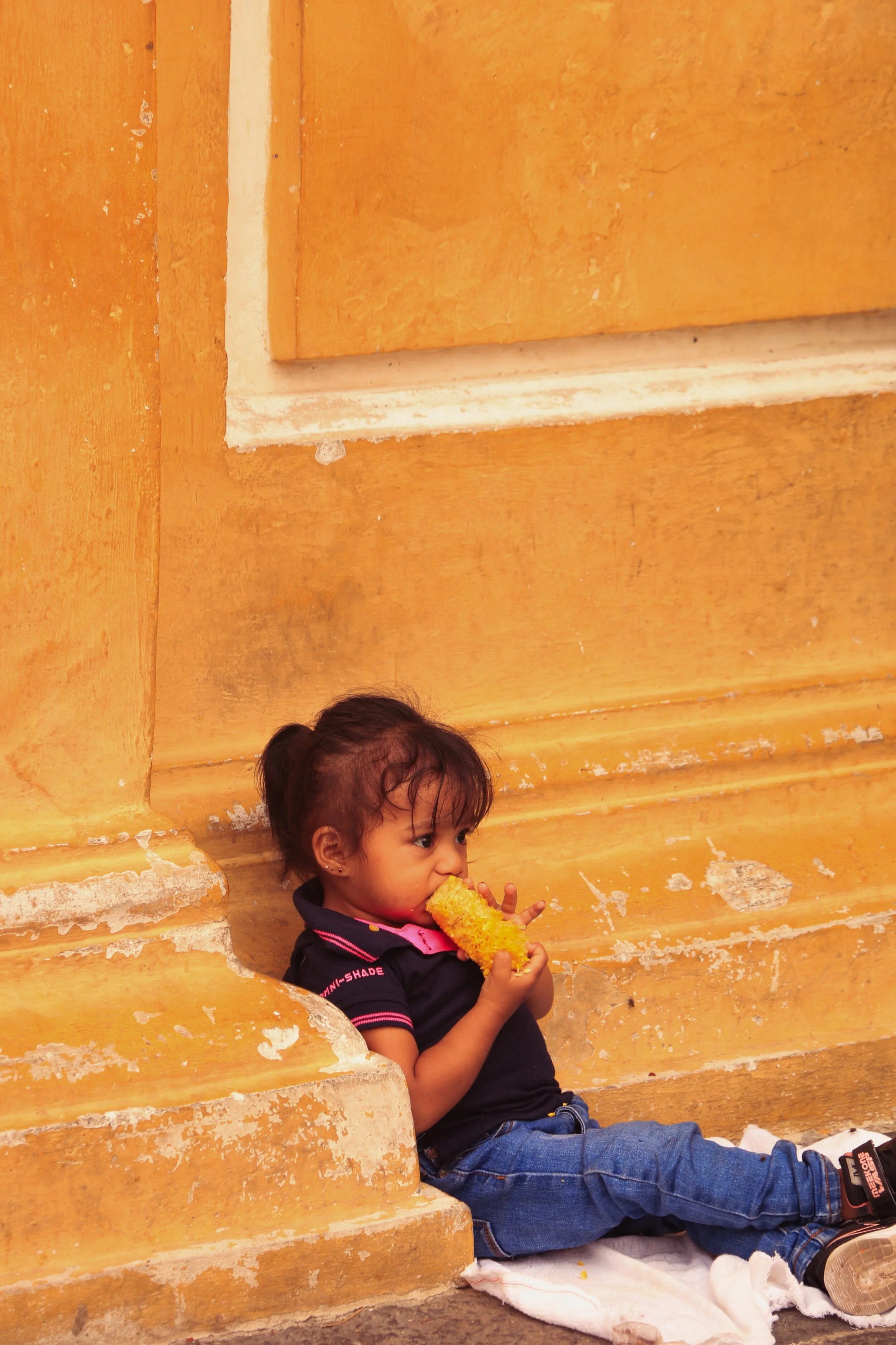 A young girl sitting on a white cloth on the ground, leaning against a yellow-painted wall, eating a corn on the cob.