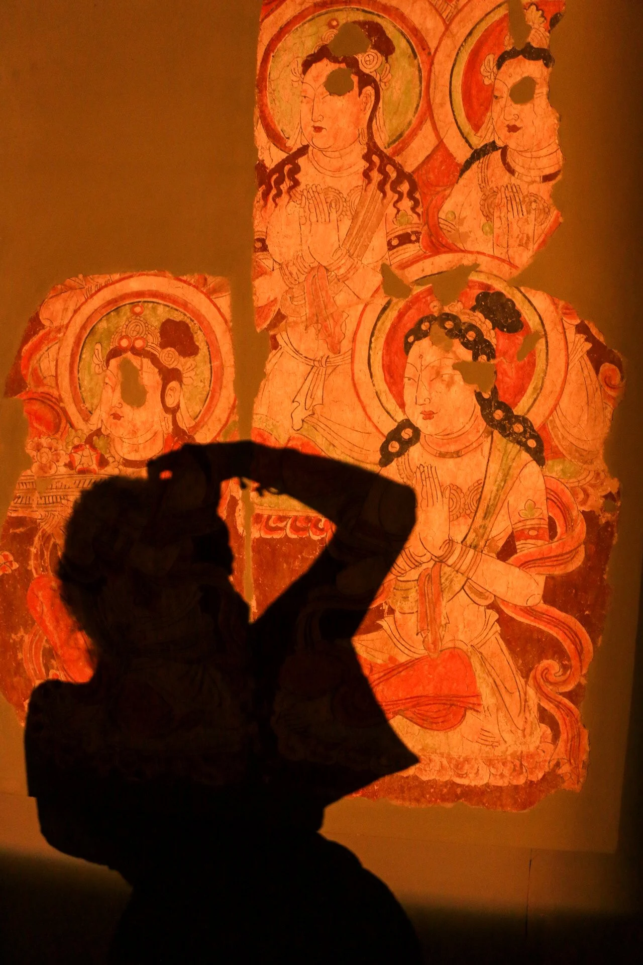 A silhouette of a person taking a photo with a smartphone in front of a brightly lit ancient mural featuring multiple deities or figures with halos, adorned with traditional clothing and jewelry.